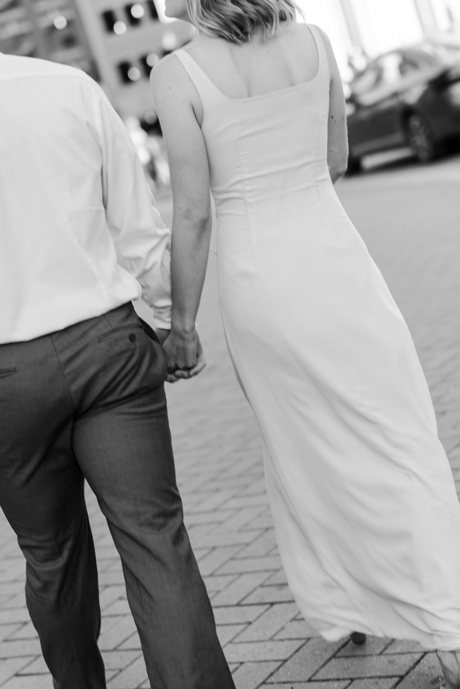 Black and white photo of a bride and groom walking hand in hand on a brick path, viewed from behind.