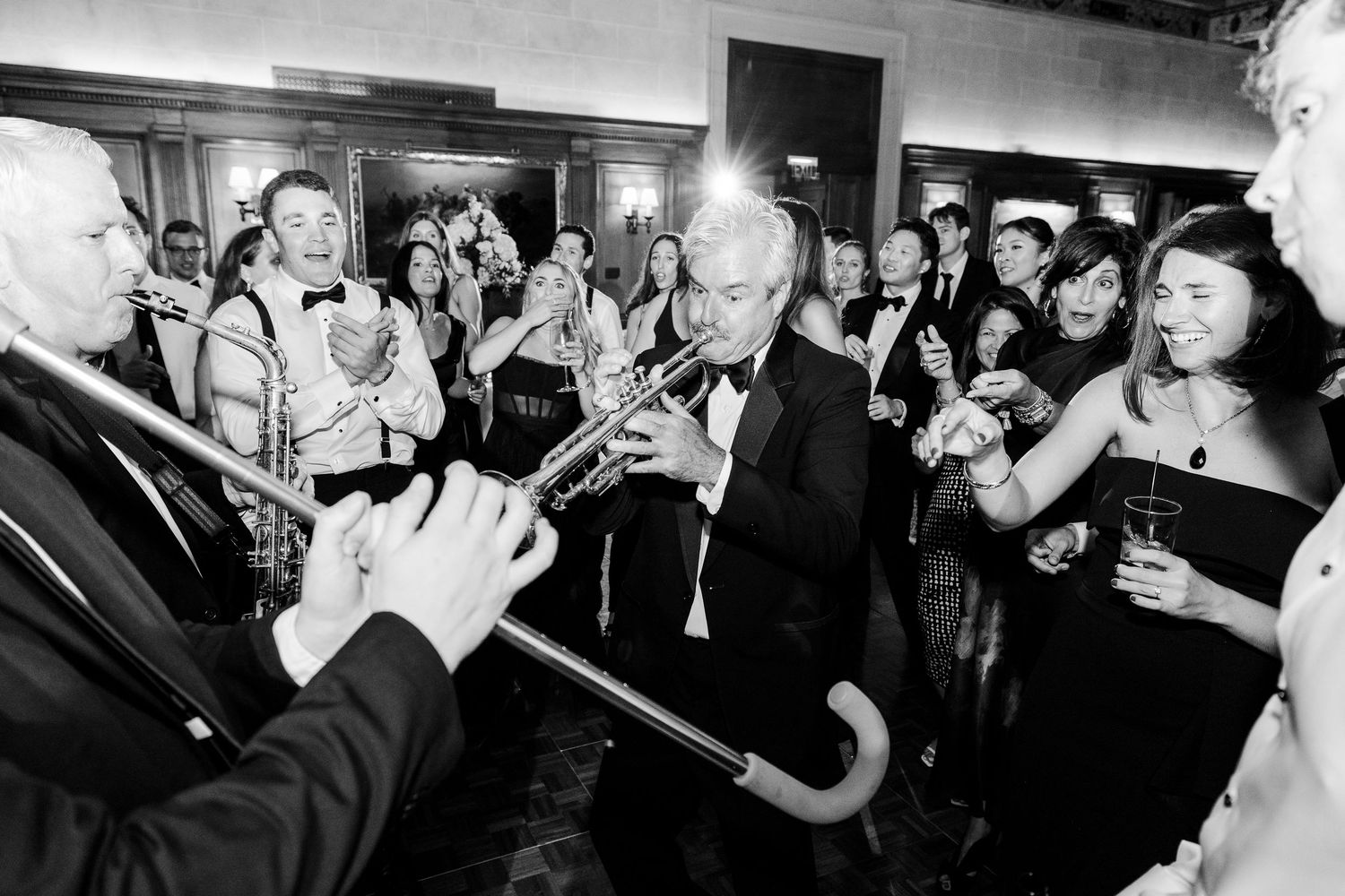 Black and white photo of a man playing trumpet at a formal event surrounded by elegantly dressed guests.