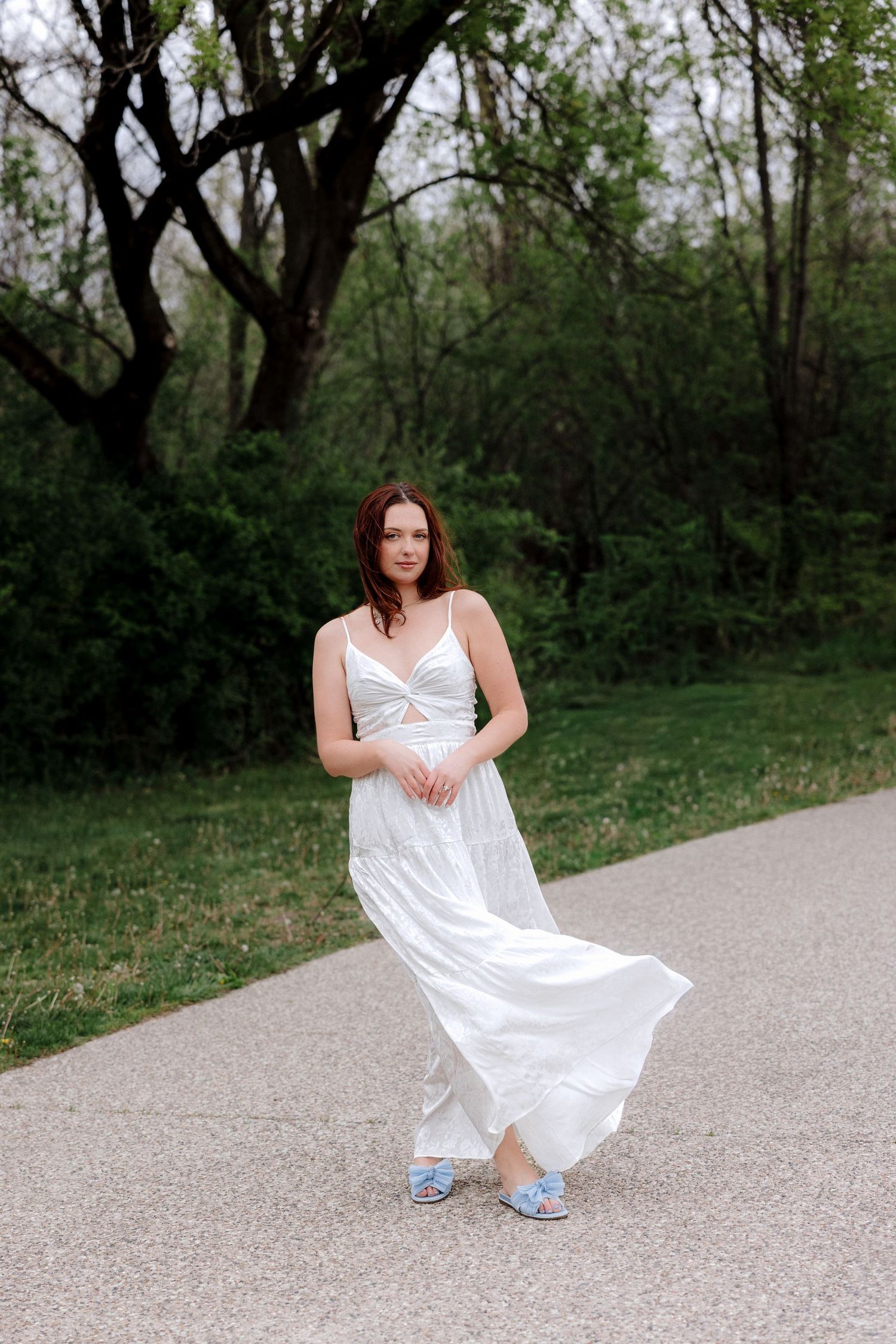 Young woman in flowing white maxi dress and blue sandals walking on a path surrounded by lush green trees.