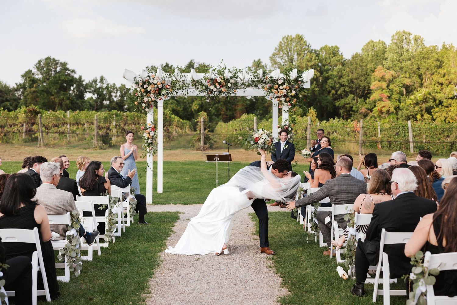 Outdoor vineyard wedding ceremony with bride and groom at floral arch, guests seated in white chairs on a lush green lawn.