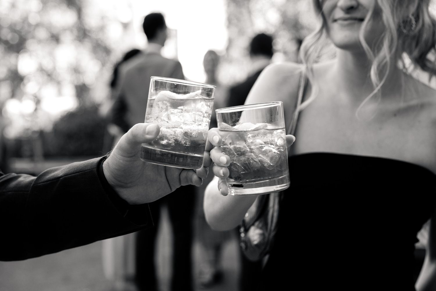 Black and white photo of two people clinking glasses of whiskey on ice at an elegant outdoor social event.