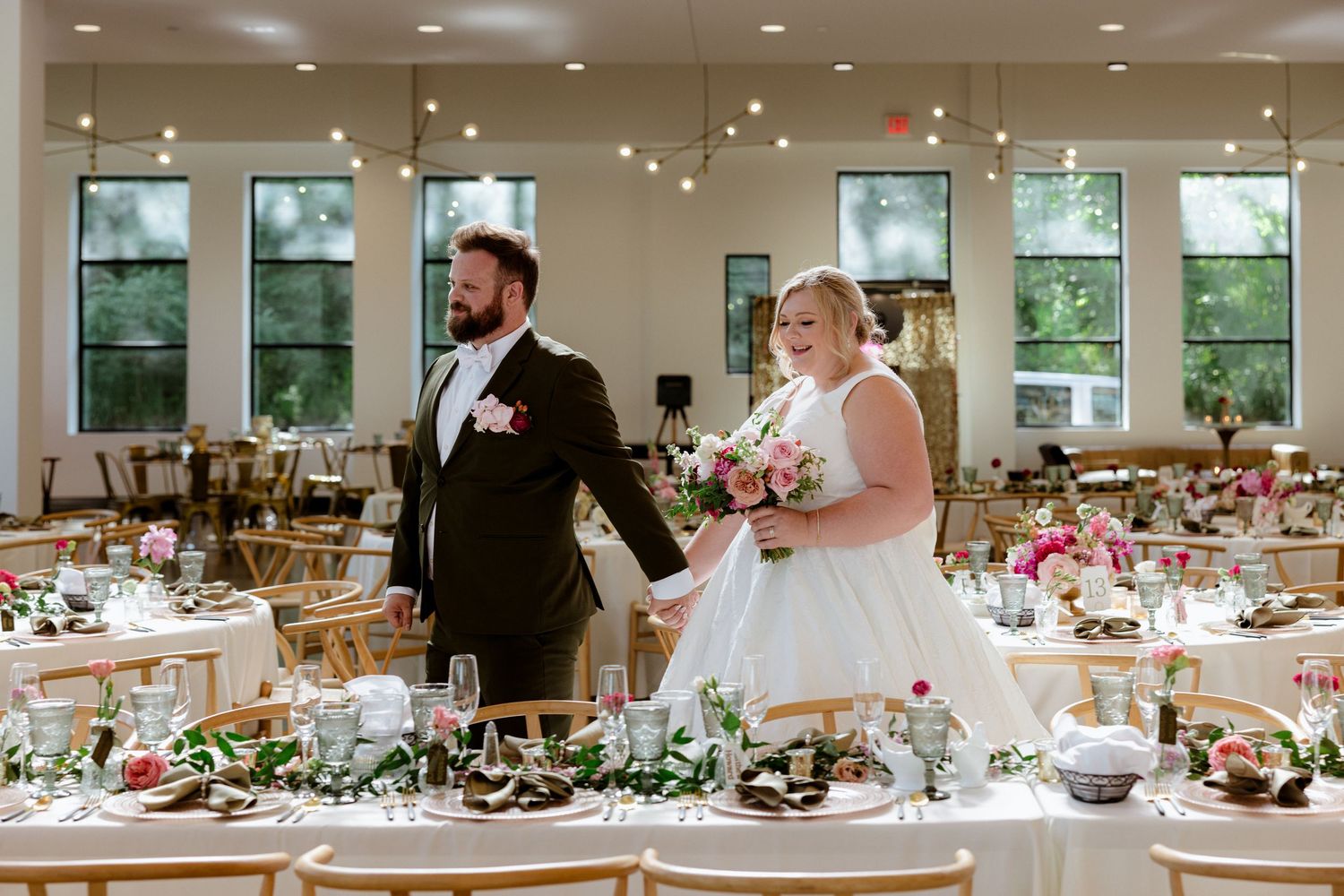 Bride and groom hold hands smiling in elegant wedding reception hall with floral centerpieces and string lights.