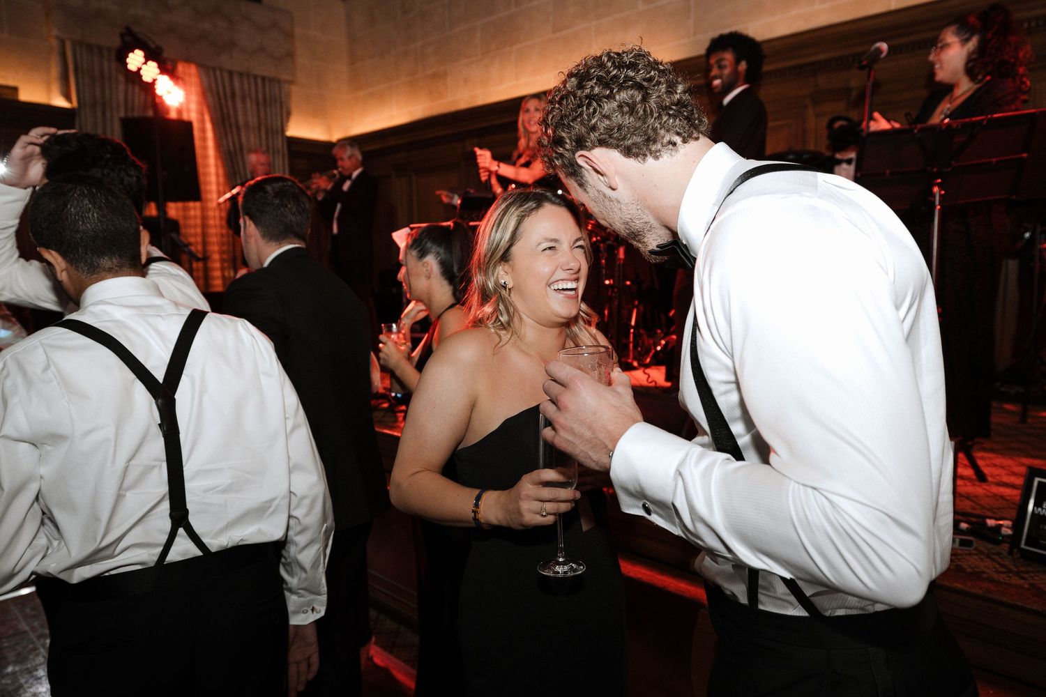 Couple in formal attire share a laugh while dancing at a warmly lit wedding reception.