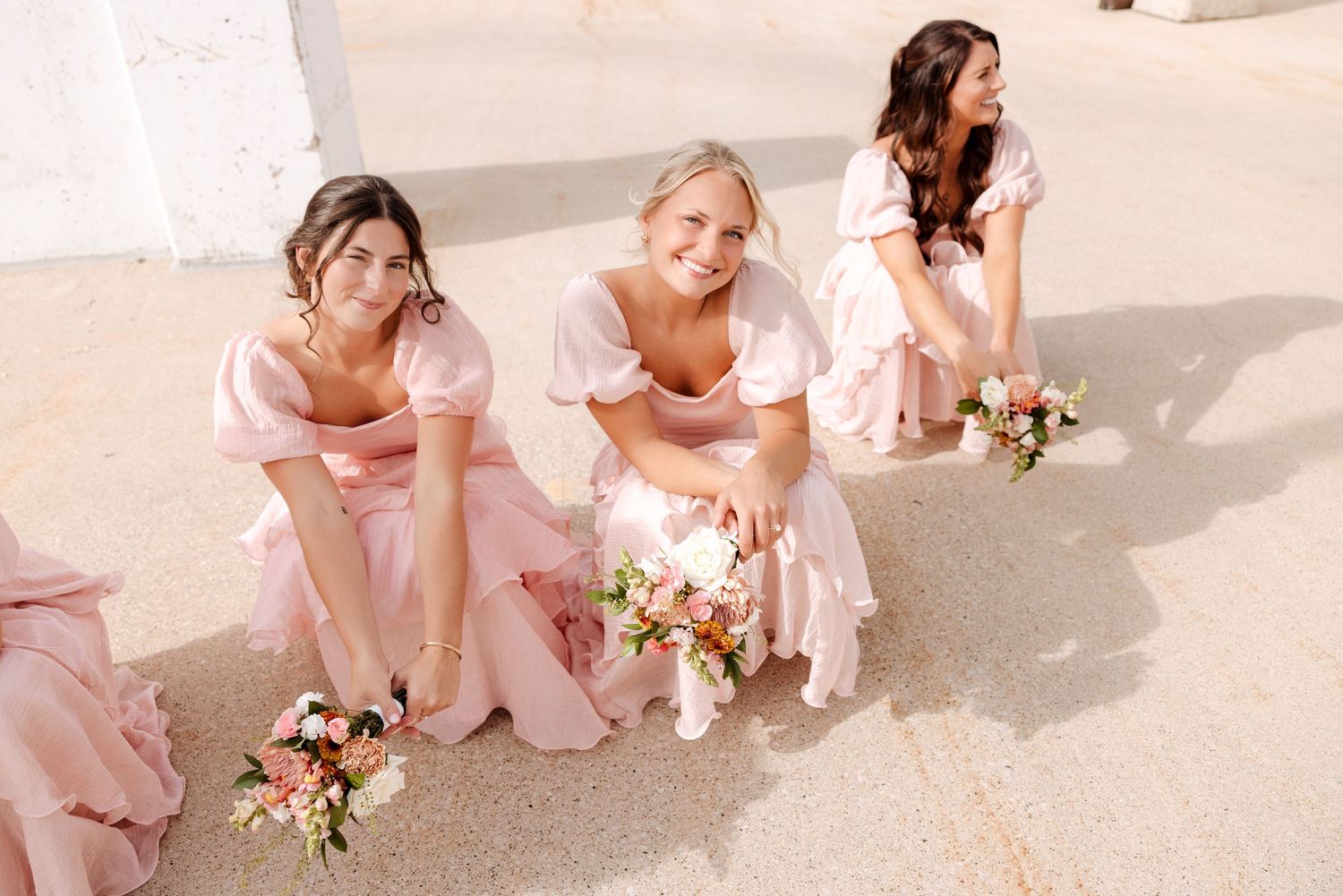 Three bridesmaids in blush pink dresses crouch together holding floral bouquets outdoors.