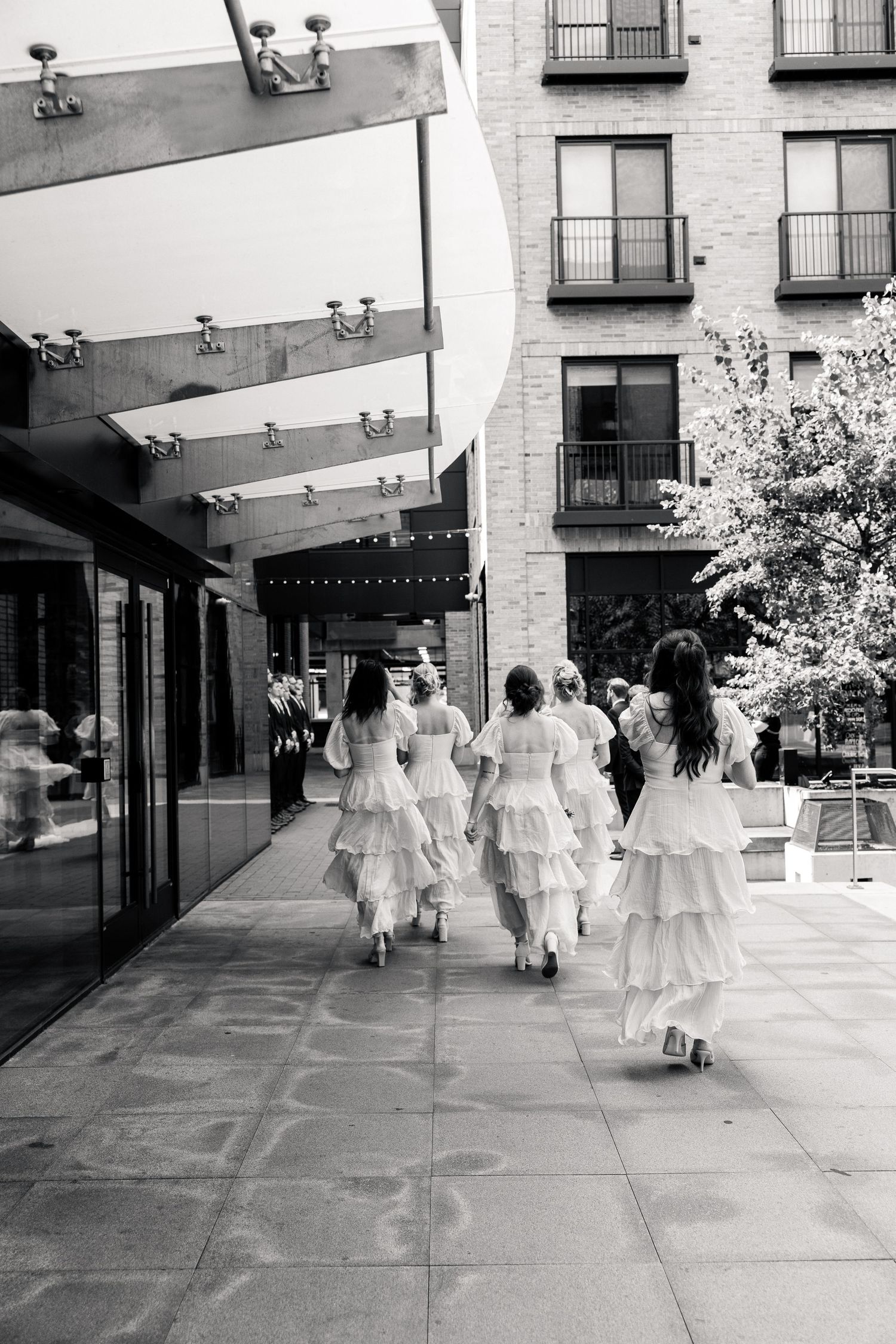 Bridesmaids in white ruffled dresses walking together along an urban sidewalk, black and white photo.