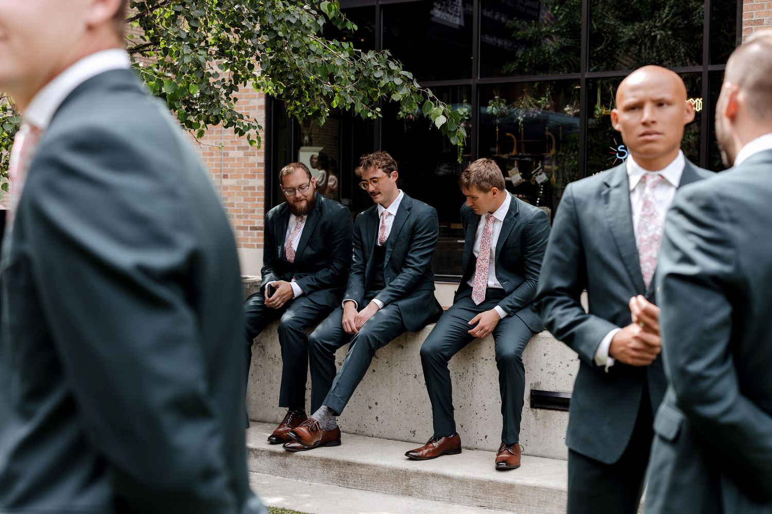 Groomsmen in dark suits and pink ties sit and stand outside during a wedding, relaxing before the ceremony.
