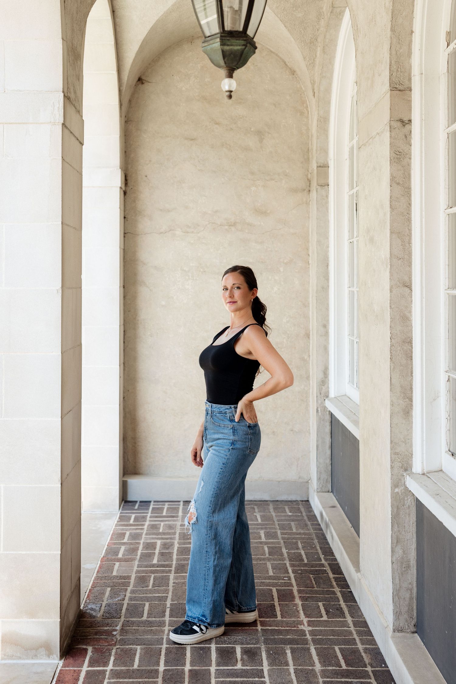 Woman in black tank top and wide-leg jeans posing in elegant arched stone corridor with brick floor.