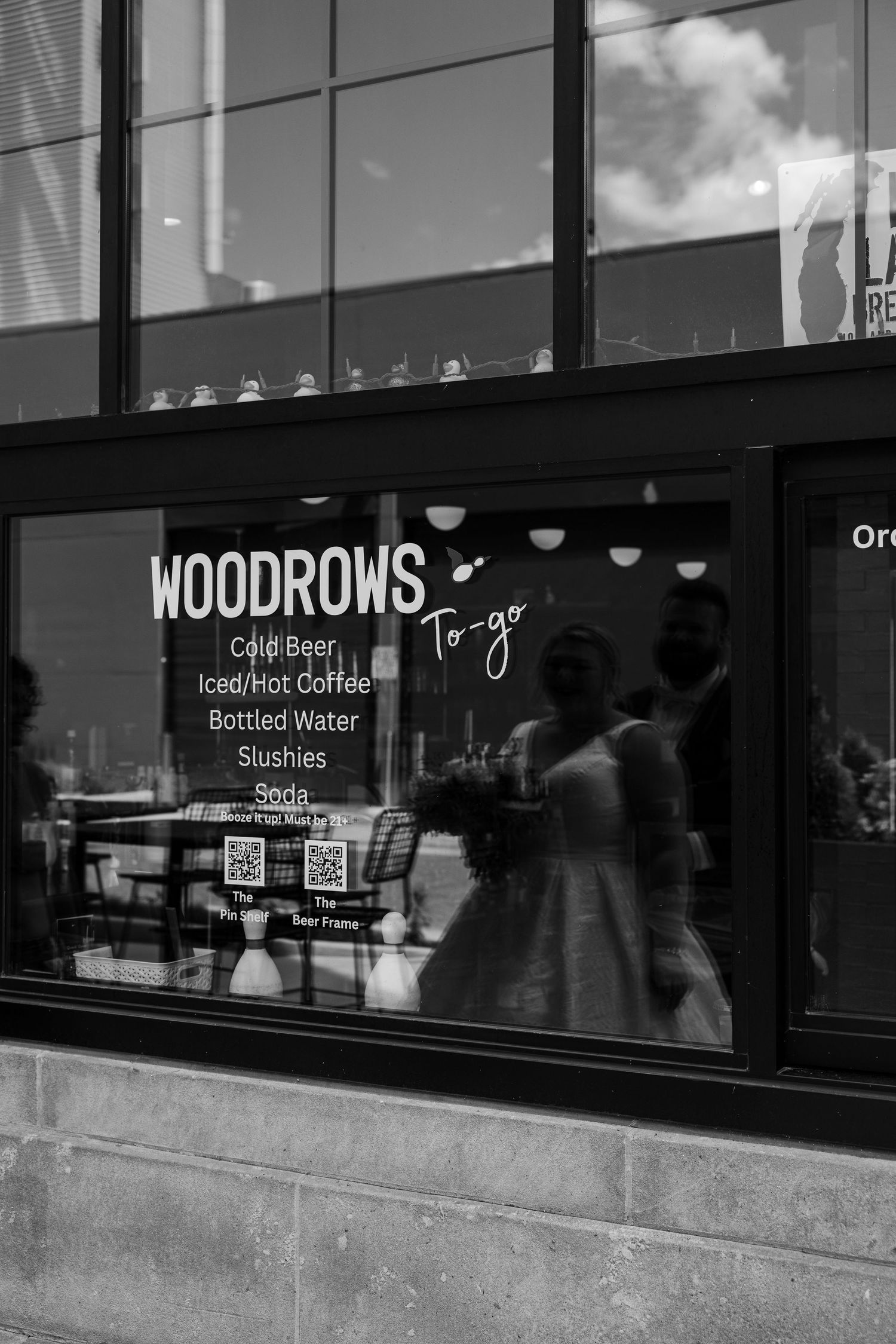 Black and white photo of Woodrows To-Go shop window displaying cold beer, coffee, slushies and soda menu.