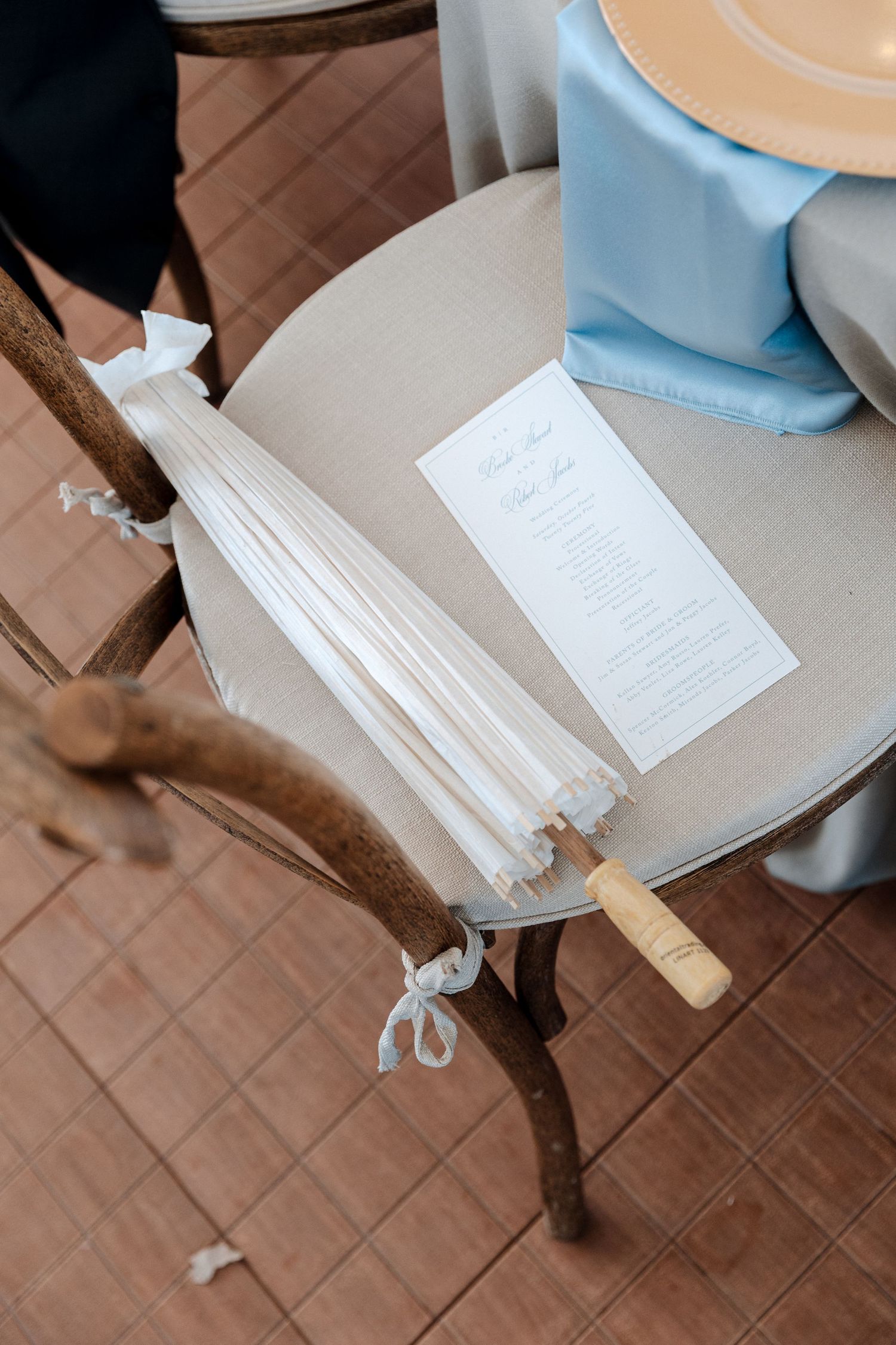 White paper fan wedding program resting on a rustic wooden chair beside a light blue tablecloth and gold plate.