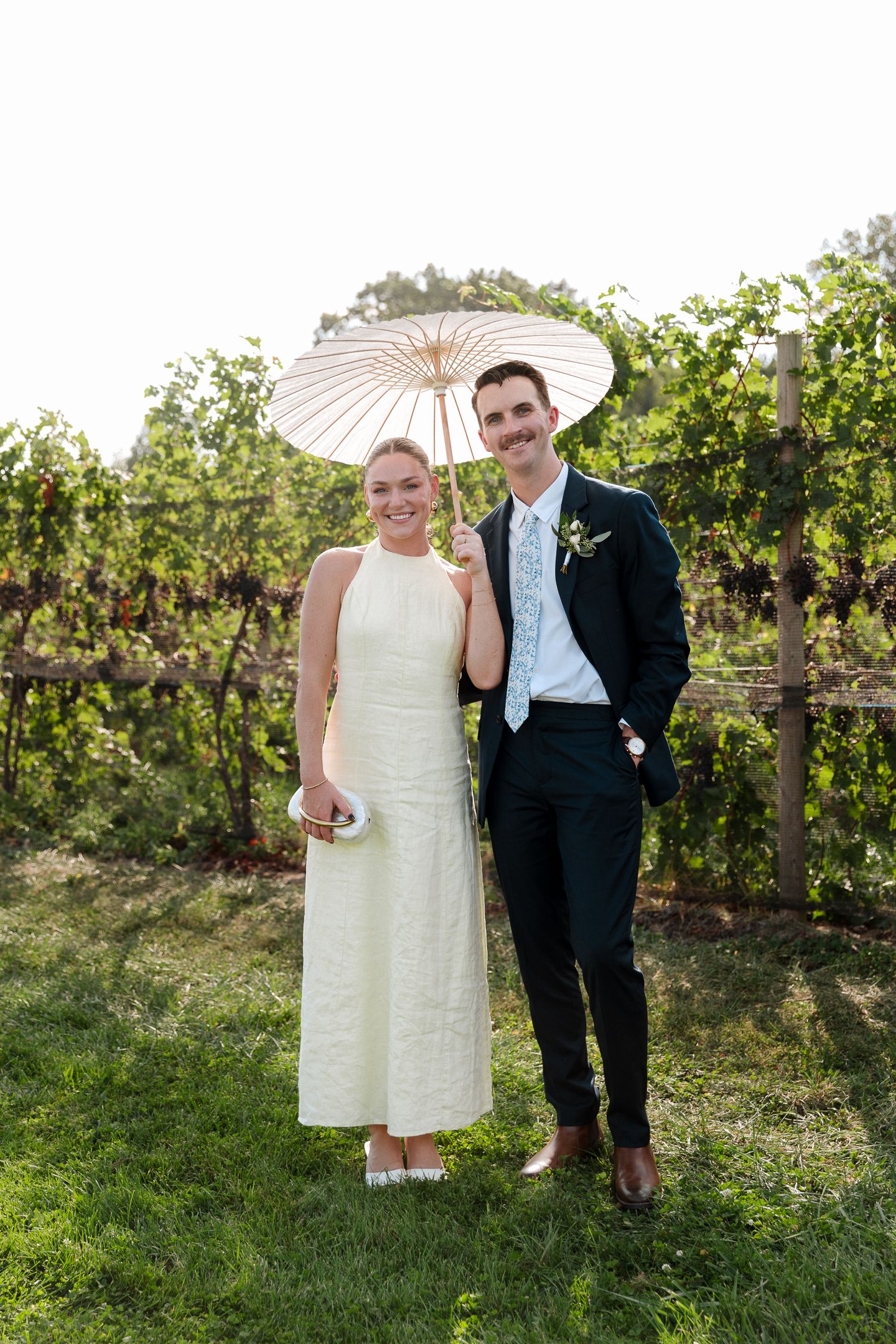 A bride in white dress and groom in dark suit pose with parasol in a lush vineyard setting.