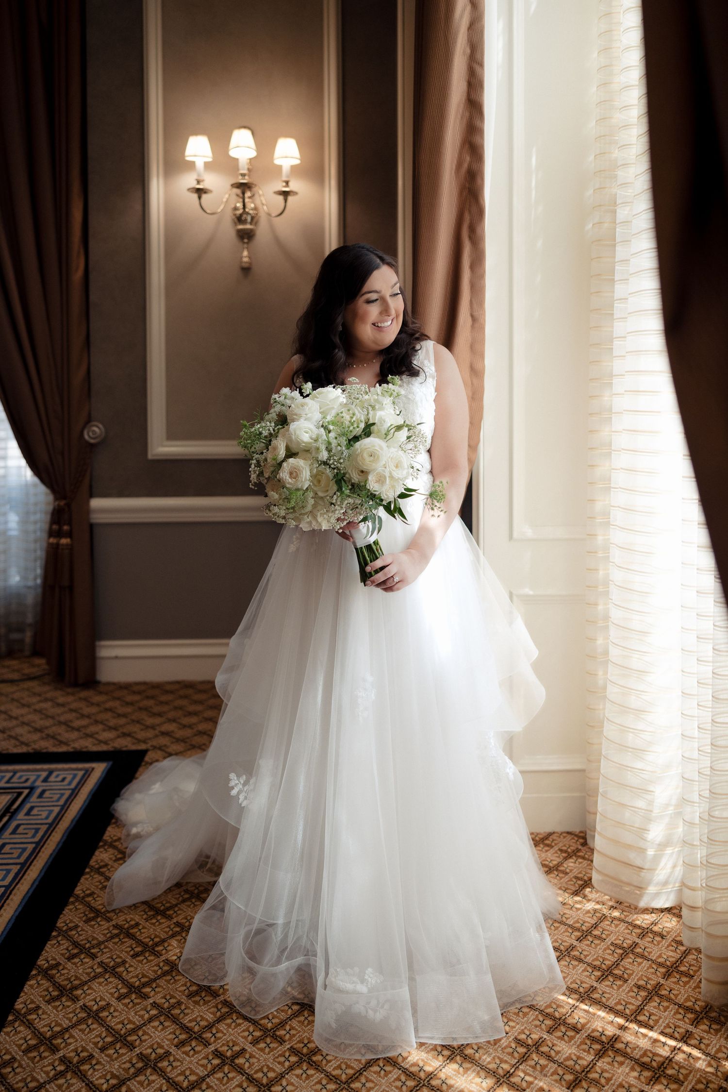 Smiling bride in layered tulle gown holds white floral bouquet by window.