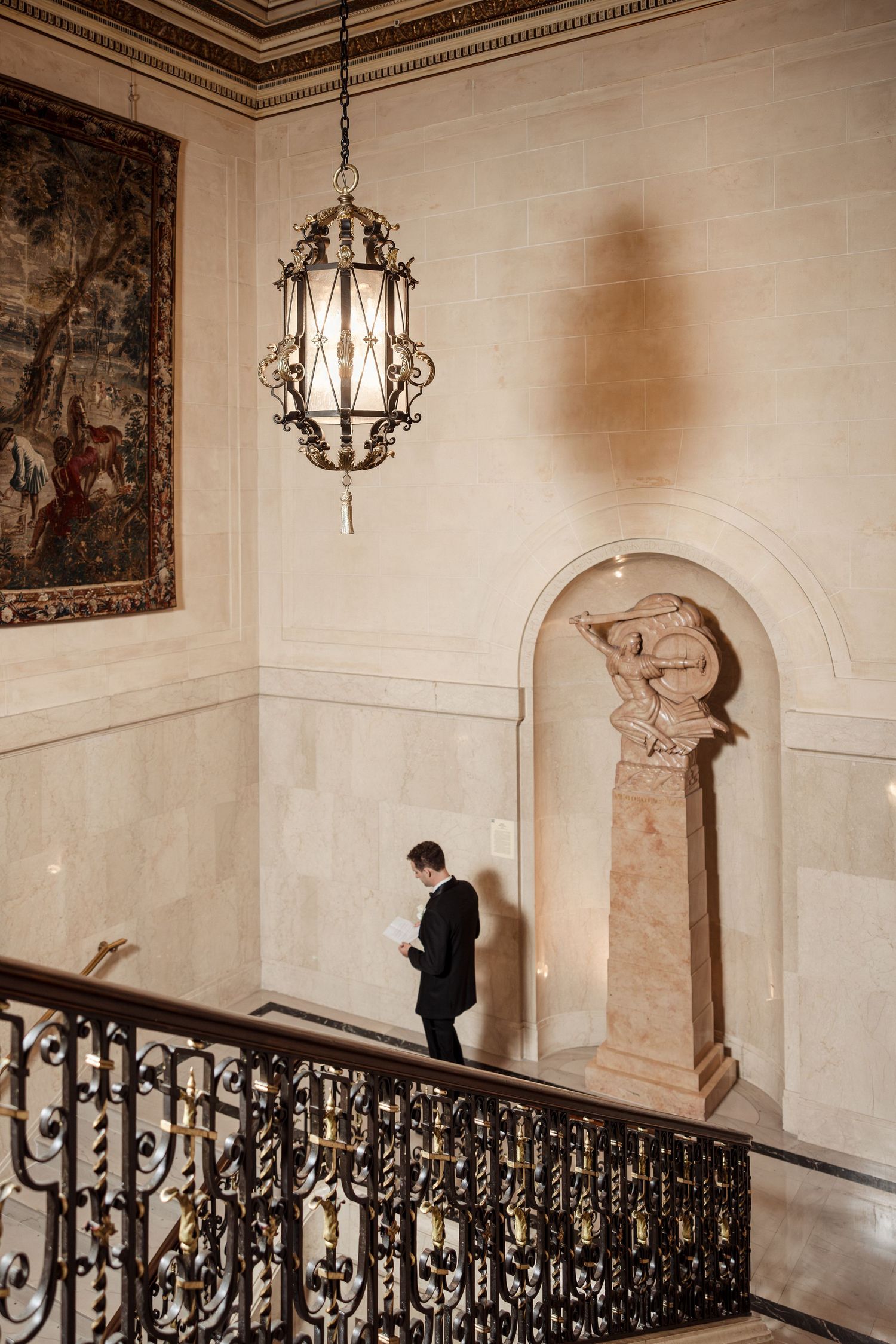 Groom in black suit walks grand staircase beneath ornate chandelier.
