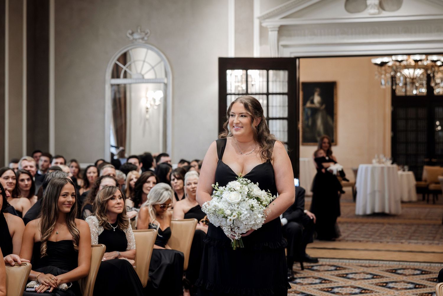 Bridesmaid in black gown walks down aisle carrying white floral bouquet at elegant wedding ceremony.