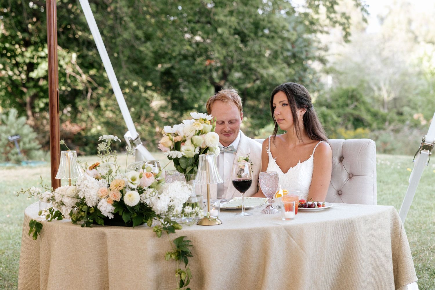 Elegant outdoor wedding reception with bride and groom seated at a floral-decorated sweetheart table with wine glasses.