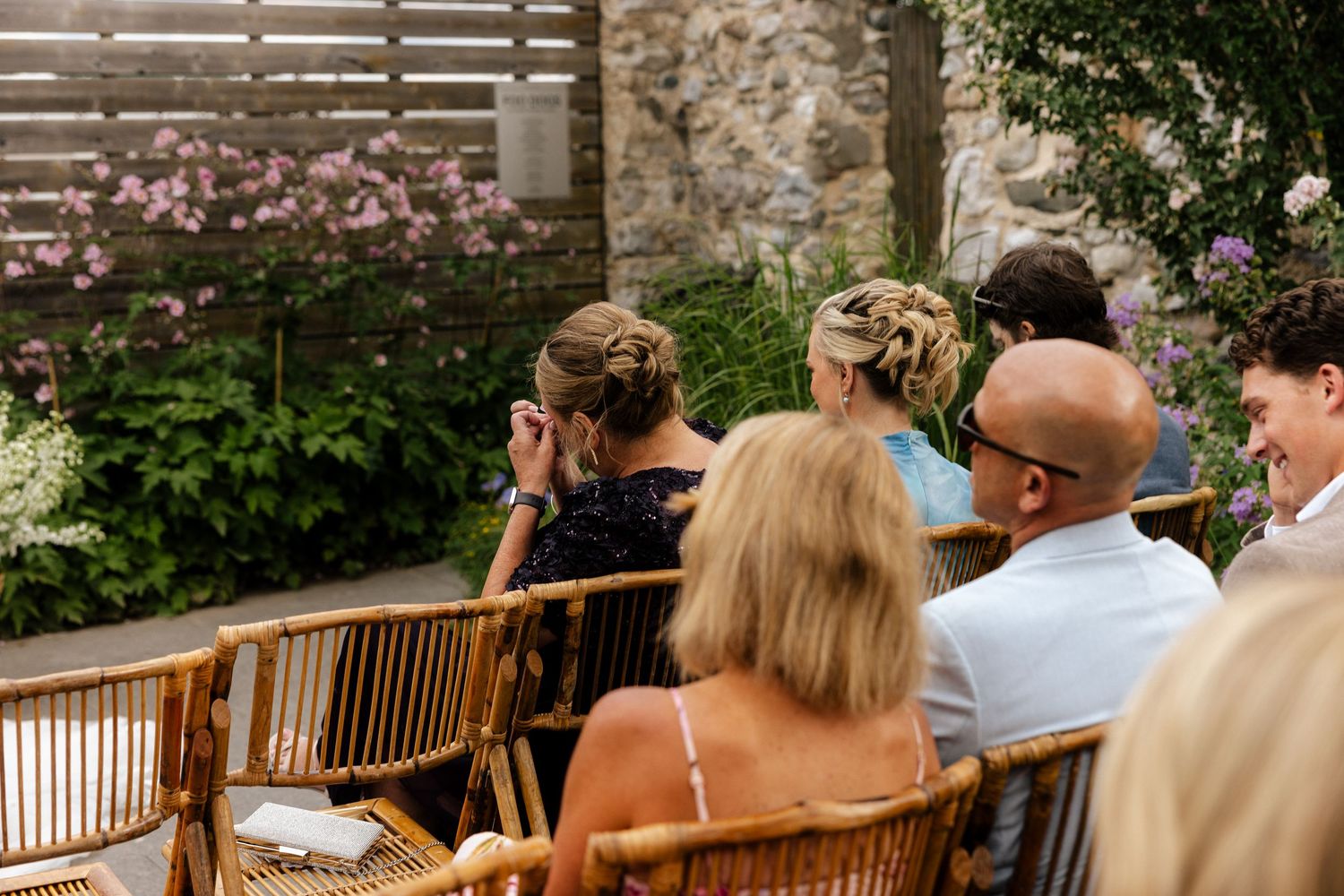 Wedding guests seated in wicker chairs outdoors, surrounded by blooming flowers and a wooden fence.