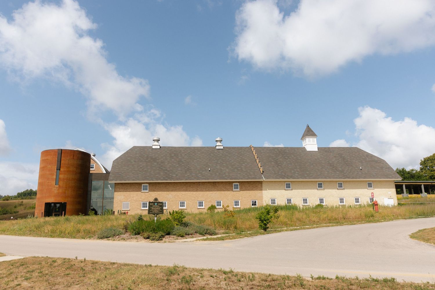 Historic barn building with cylindrical silo, white cupola, and brick facade under a partly cloudy blue sky.
