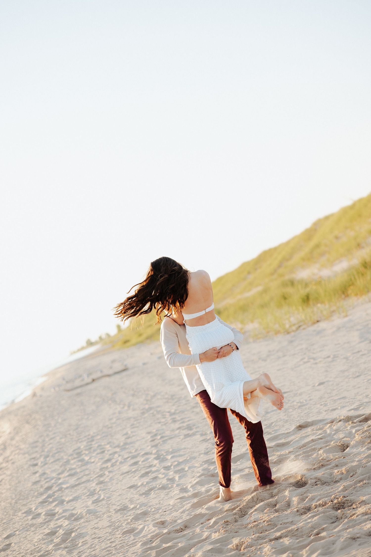 Couple embracing and spinning on a sandy beach near grass dunes in warm golden sunlight.