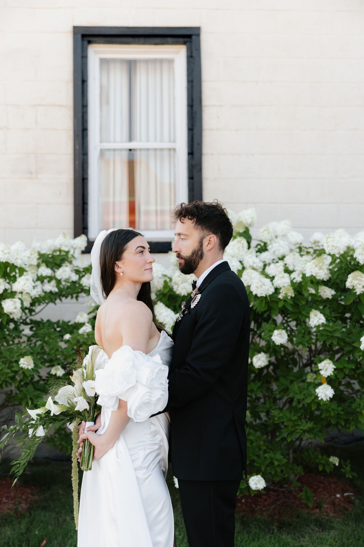 Bride and groom pose together in front of white hydrangeas, she wears an off-shoulder gown holding white florals.