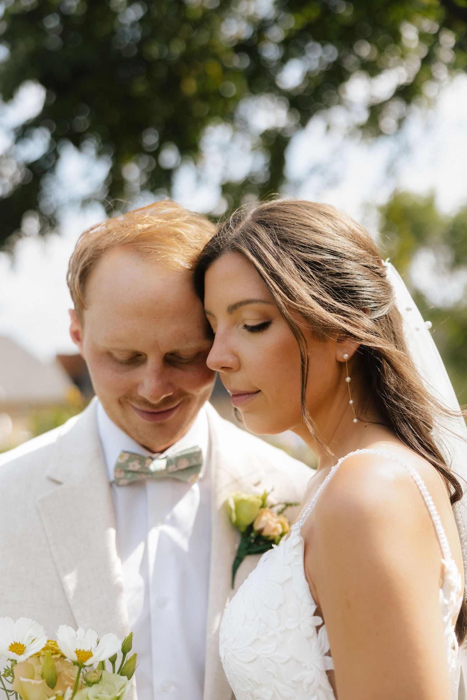 Bride and groom share a tender moment outdoors, with the bride in lace dress and groom in white suit with green bow tie.