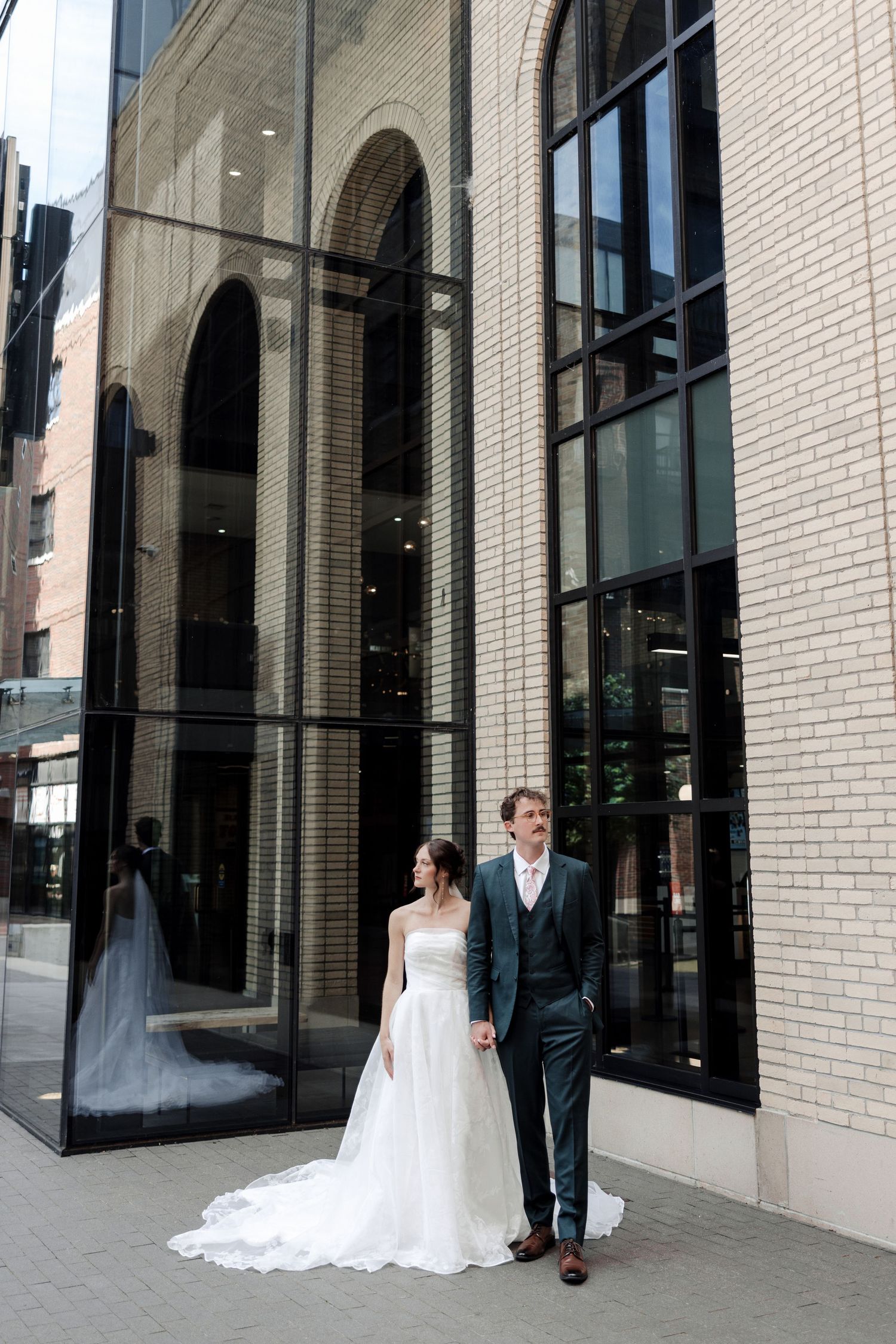 Bride in a flowing white gown and groom in a gray suit hold hands before a grand modern building with arched windows.