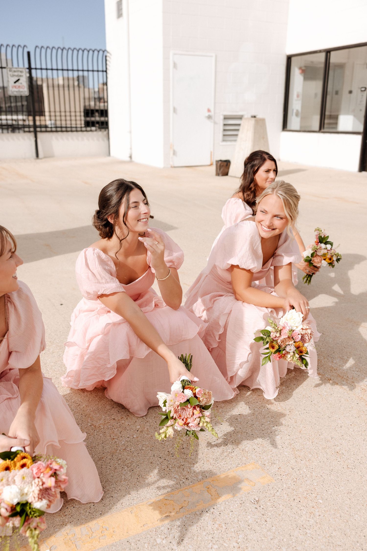 Bridesmaids in pink ruffled dresses sit laughing outdoors holding colorful floral bouquets on rooftop.