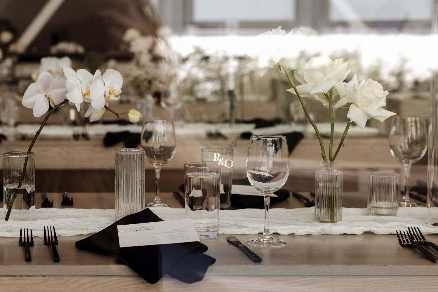 Elegant wedding reception table with white orchids, tulips, crystal glassware, and navy napkins on a rustic wooden table.