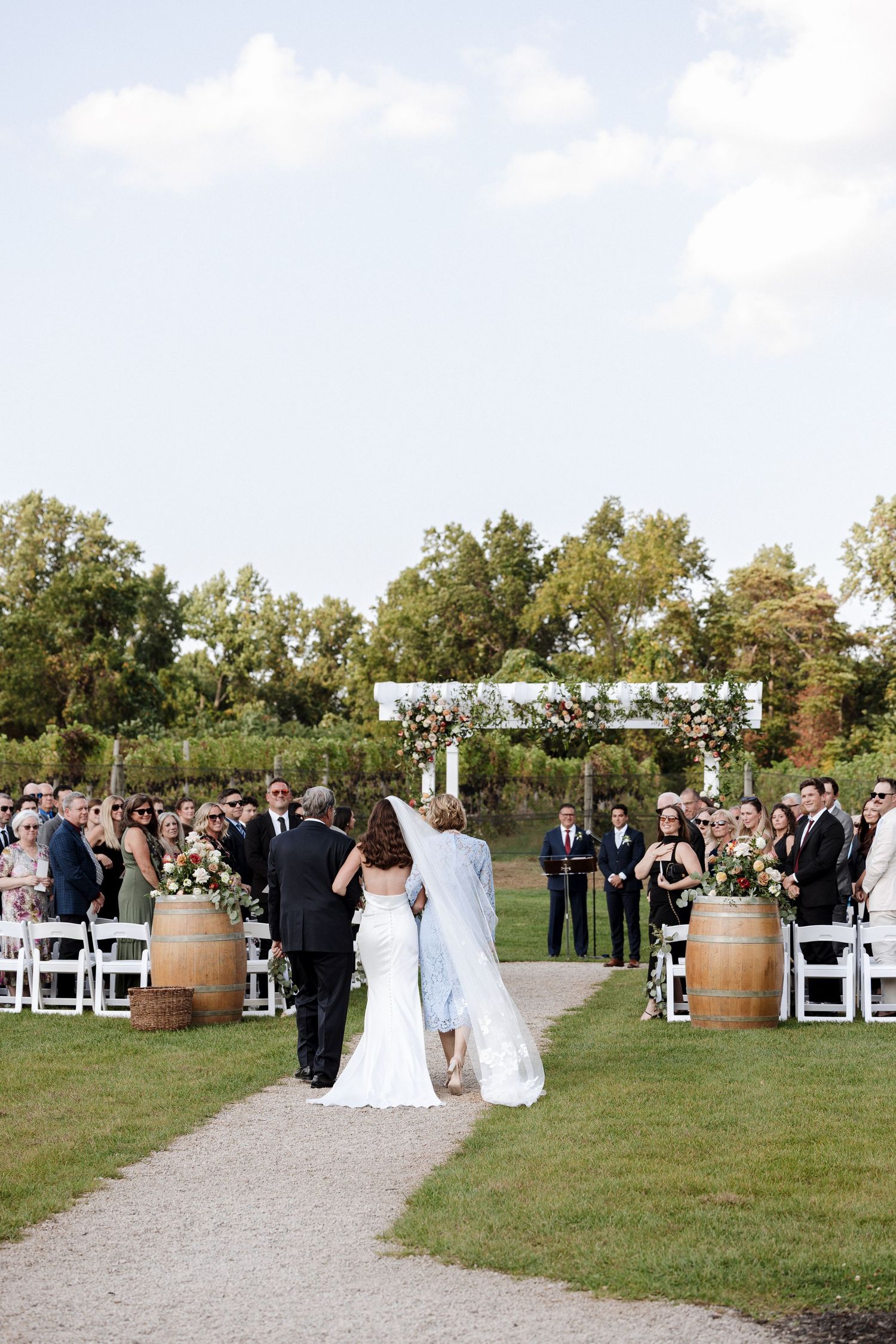 Outdoor vineyard wedding ceremony with bride walking down gravel aisle flanked by barrel arrangements toward floral arch.
