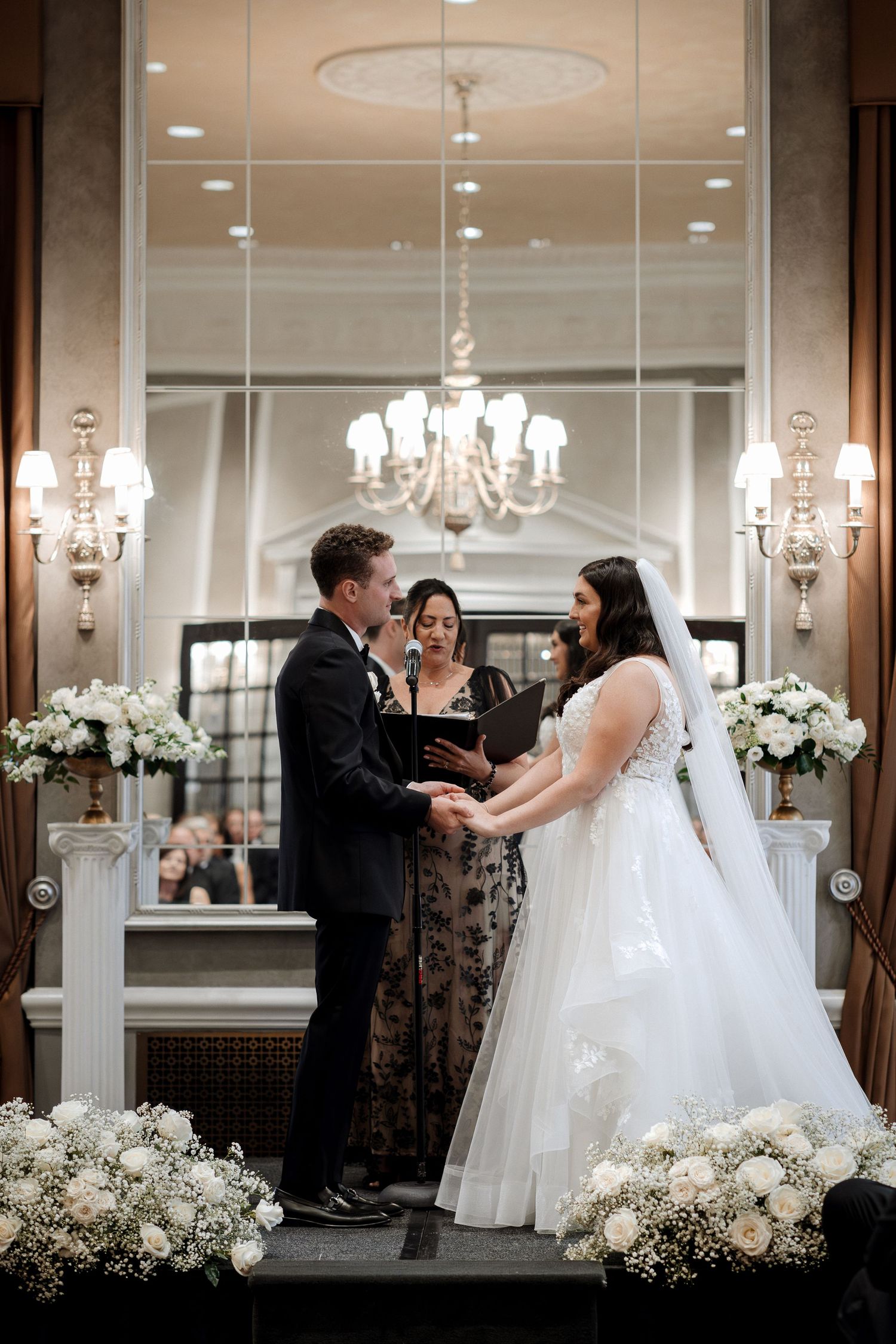 Couple holds hands exchanging vows with officiant before a grand mirror and white floral arrangements at altar.