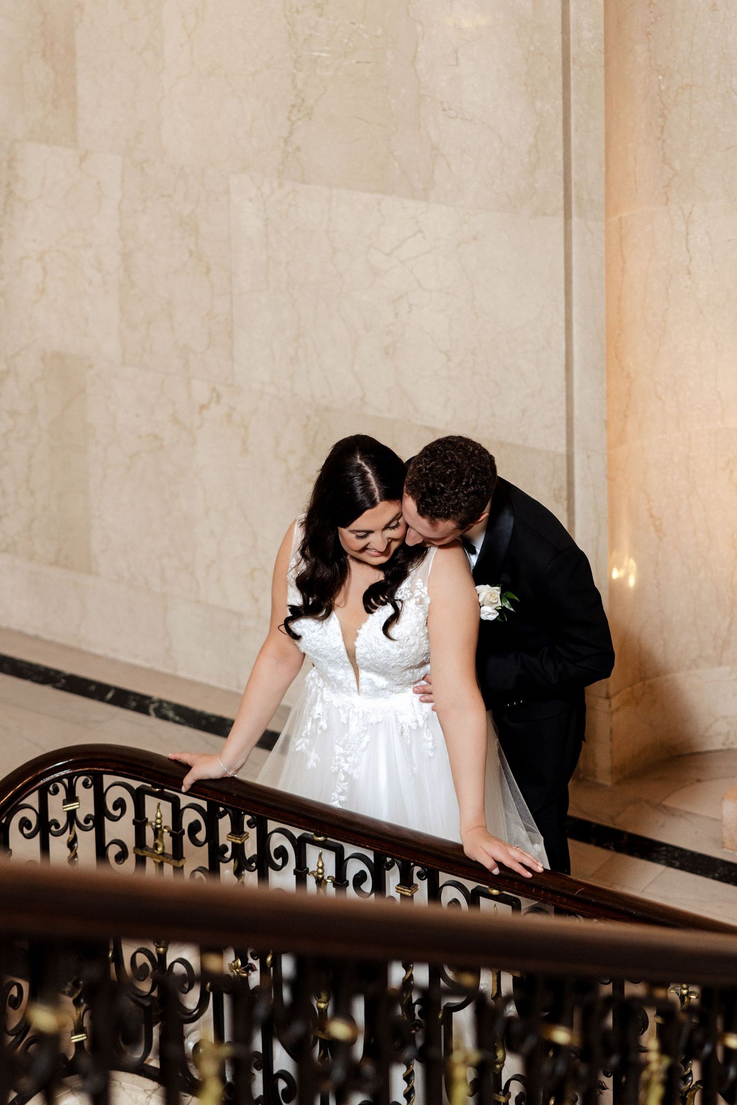 Groom tenderly kisses bride on ornate marble staircase with decorative iron railing at elegant venue.