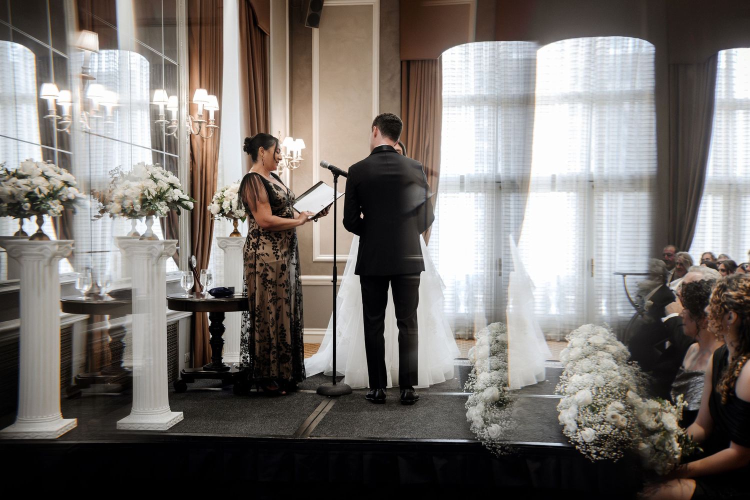 Officiant in a gold gown leads wedding ceremony as groom stands at altar flanked by white floral arrangements.