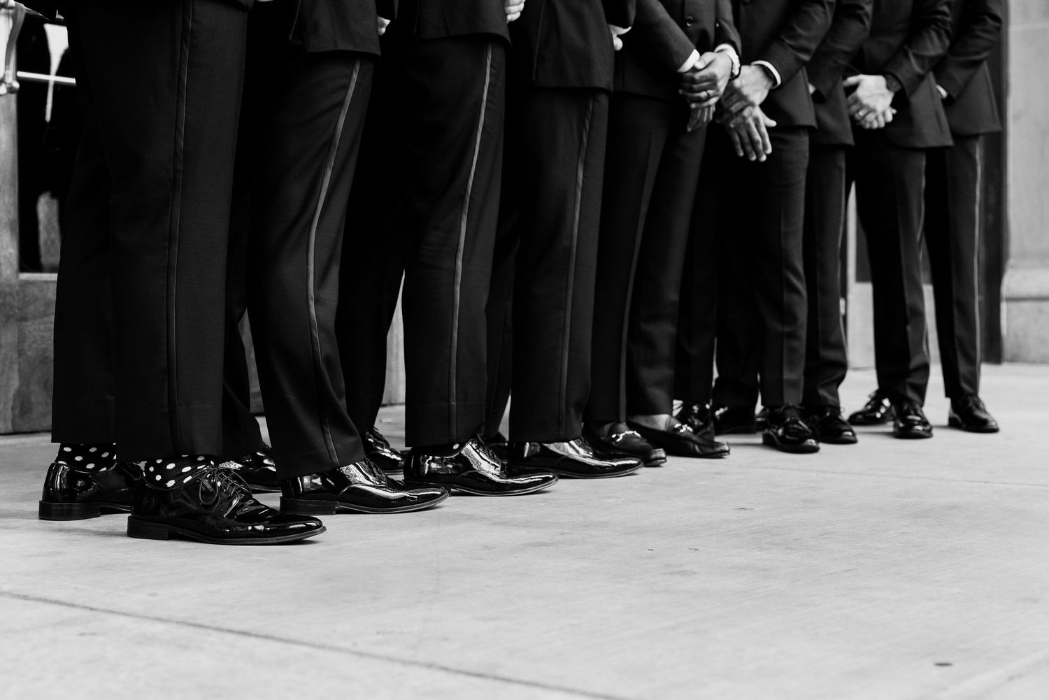 Black and white photo of groomsmen in suits showing polished dress shoes and patterned socks lined up at a wedding.