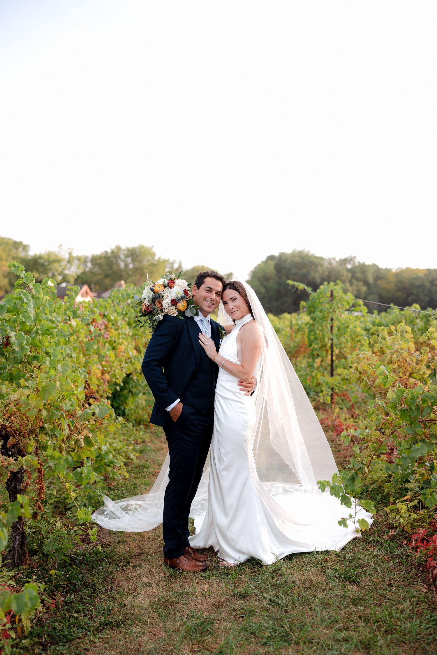 Bride and groom embracing in a vineyard, bride wearing white gown with veil holding bouquet, groom in navy suit.
