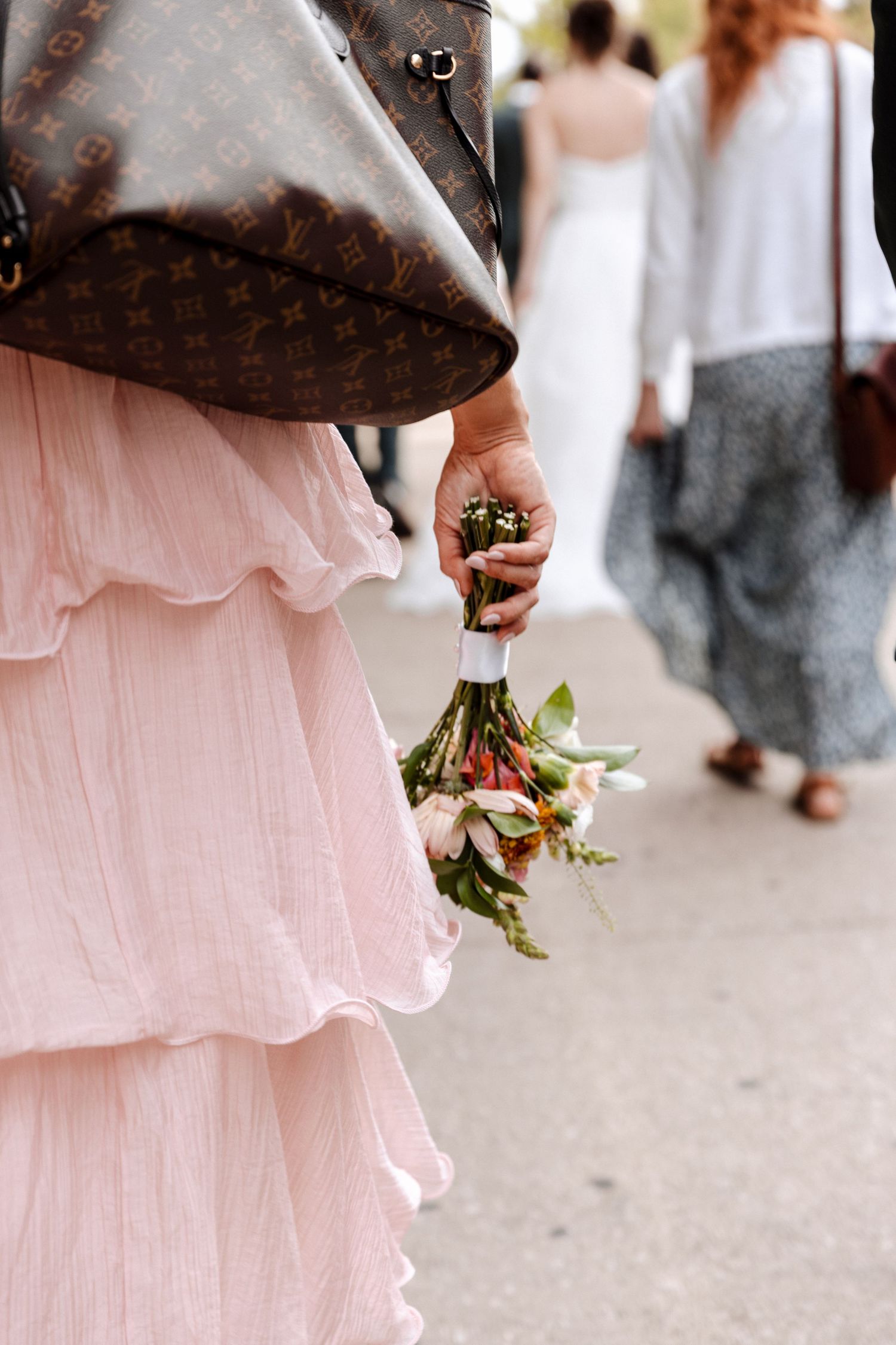 Bridesmaid in a pink ruffled dress holds a small floral bouquet while wearing a Louis Vuitton jacket.