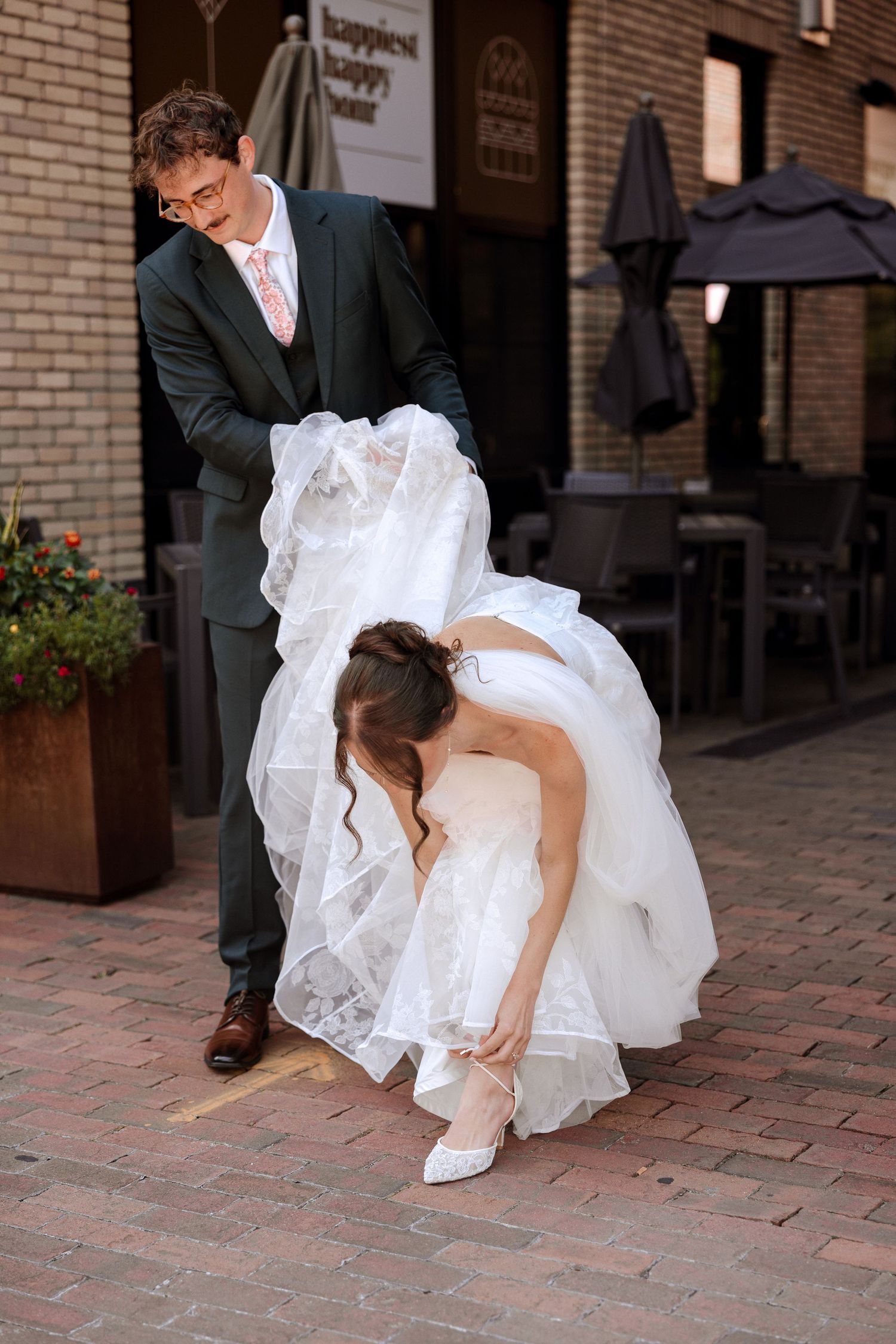 Groom lifts bride's white wedding gown as she adjusts her white heels on a brick sidewalk outside a venue.