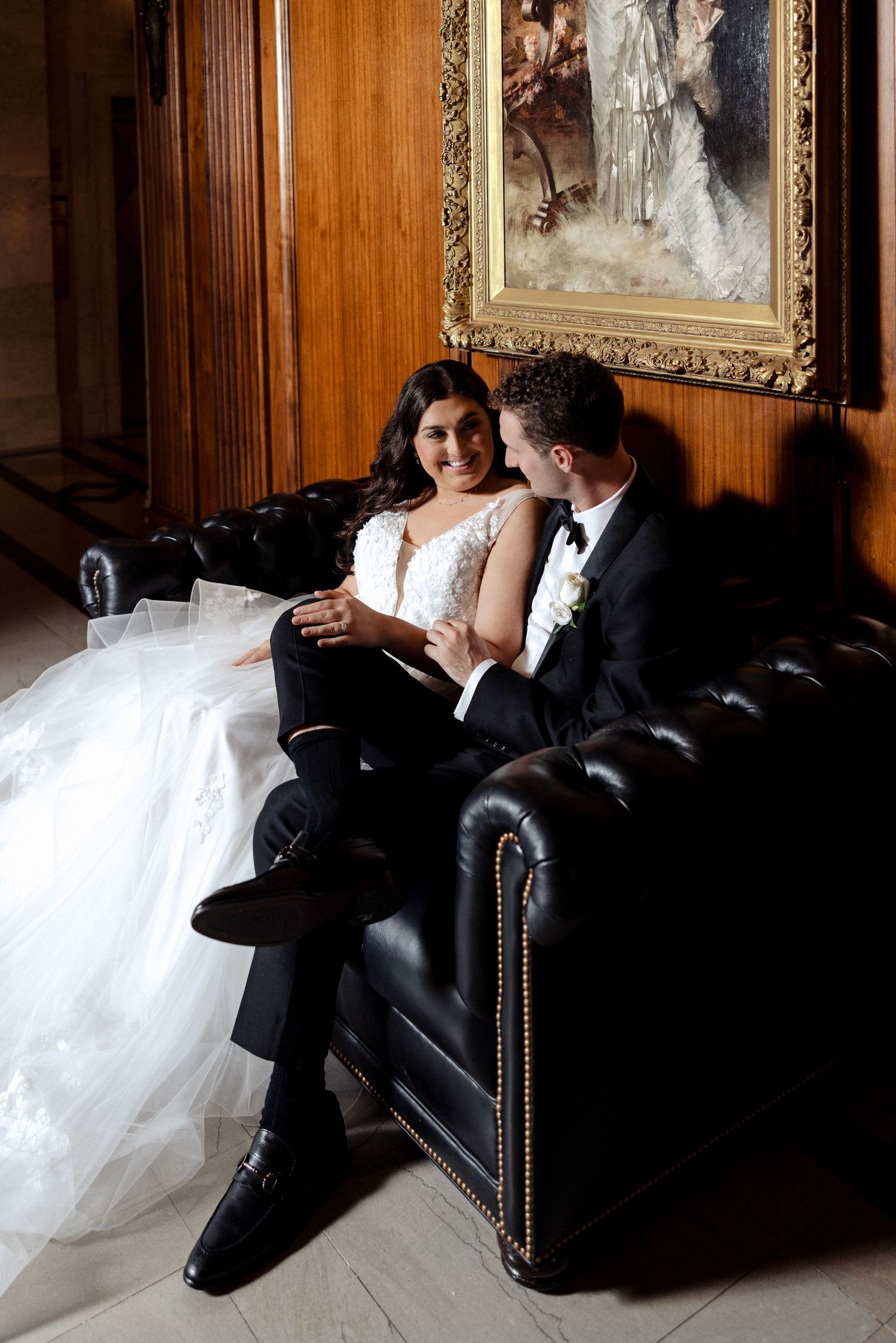 Smiling bride and groom relaxing together on a leather sofa beneath a classical oil painting.
