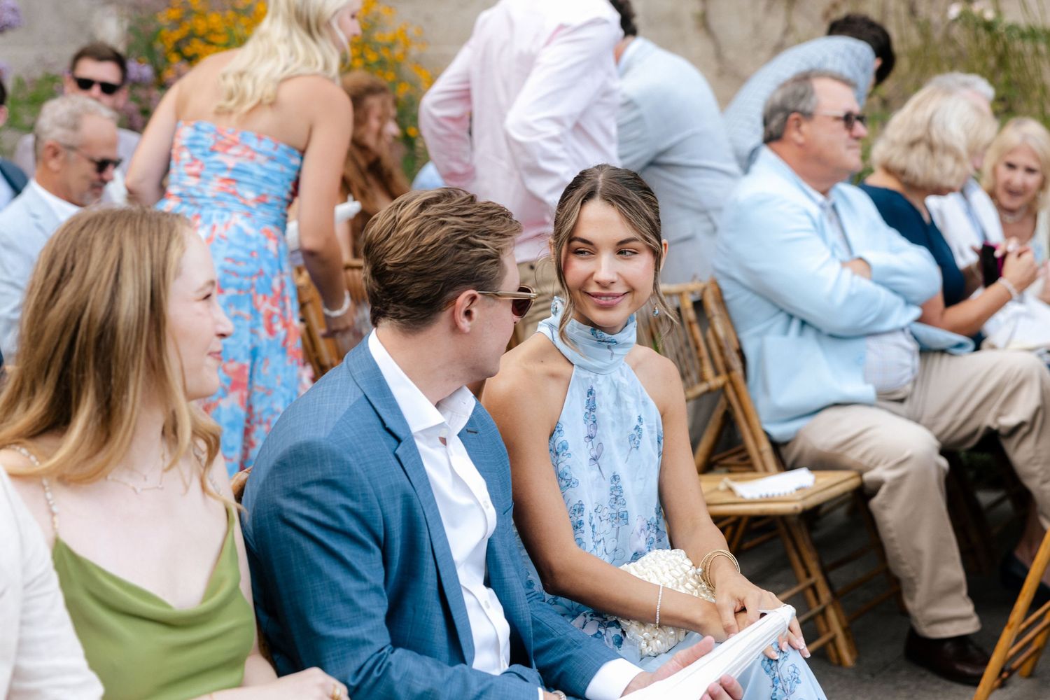 Guests seated outdoors at a wedding ceremony, with a woman in a blue floral dress smiling.