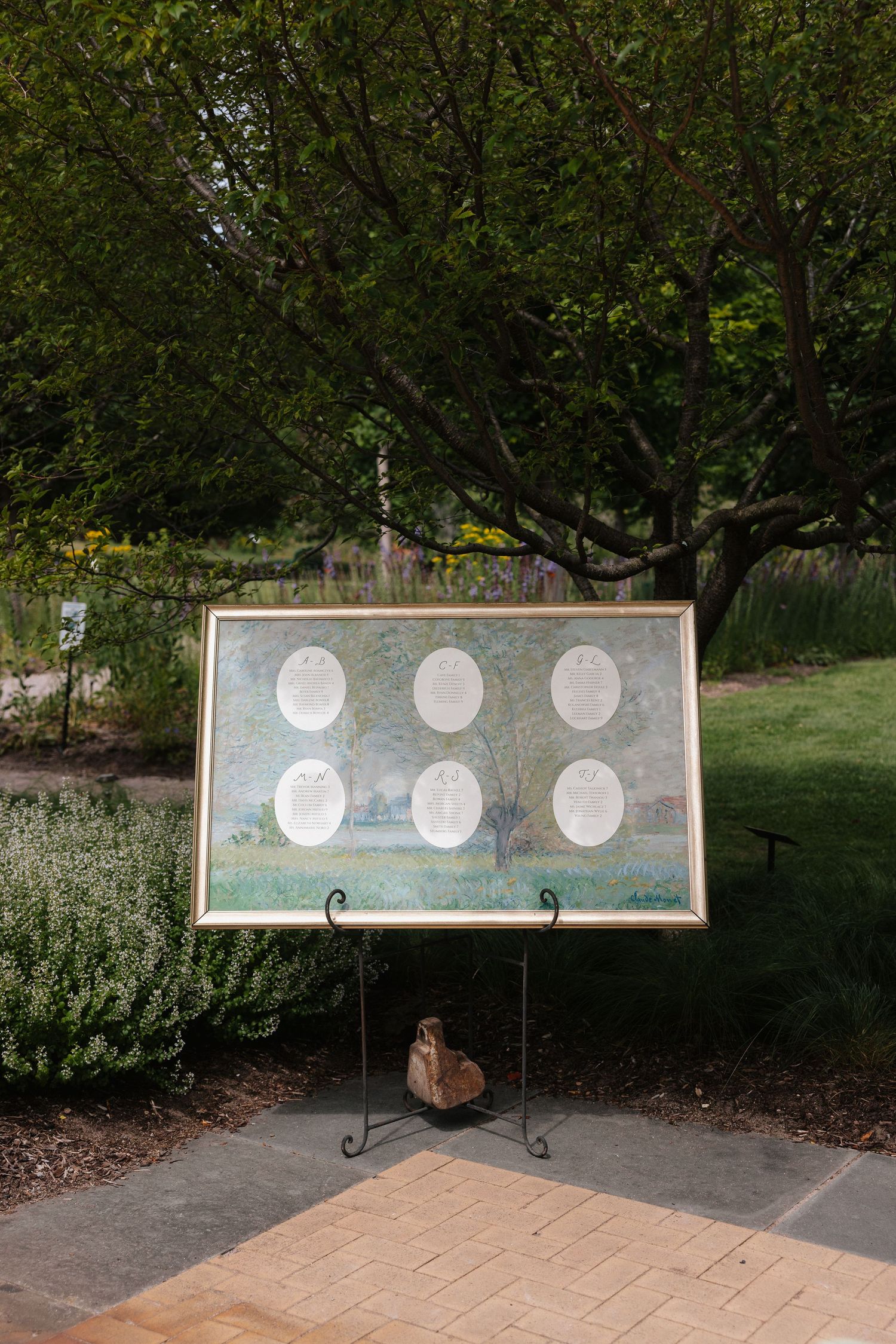 Outdoor wedding seating chart displayed on a framed painting on an easel, surrounded by lush greenery.