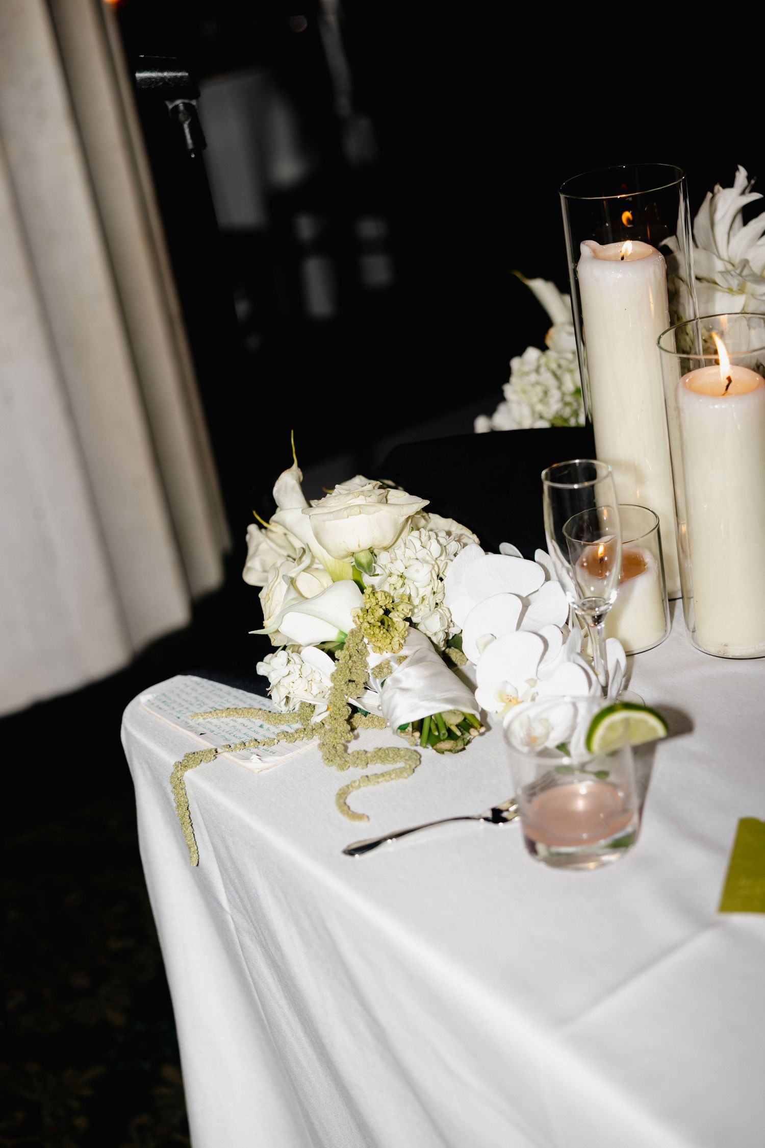 Elegant wedding table with white floral bouquet, lit pillar candles, and white linen tablecloth at a reception.