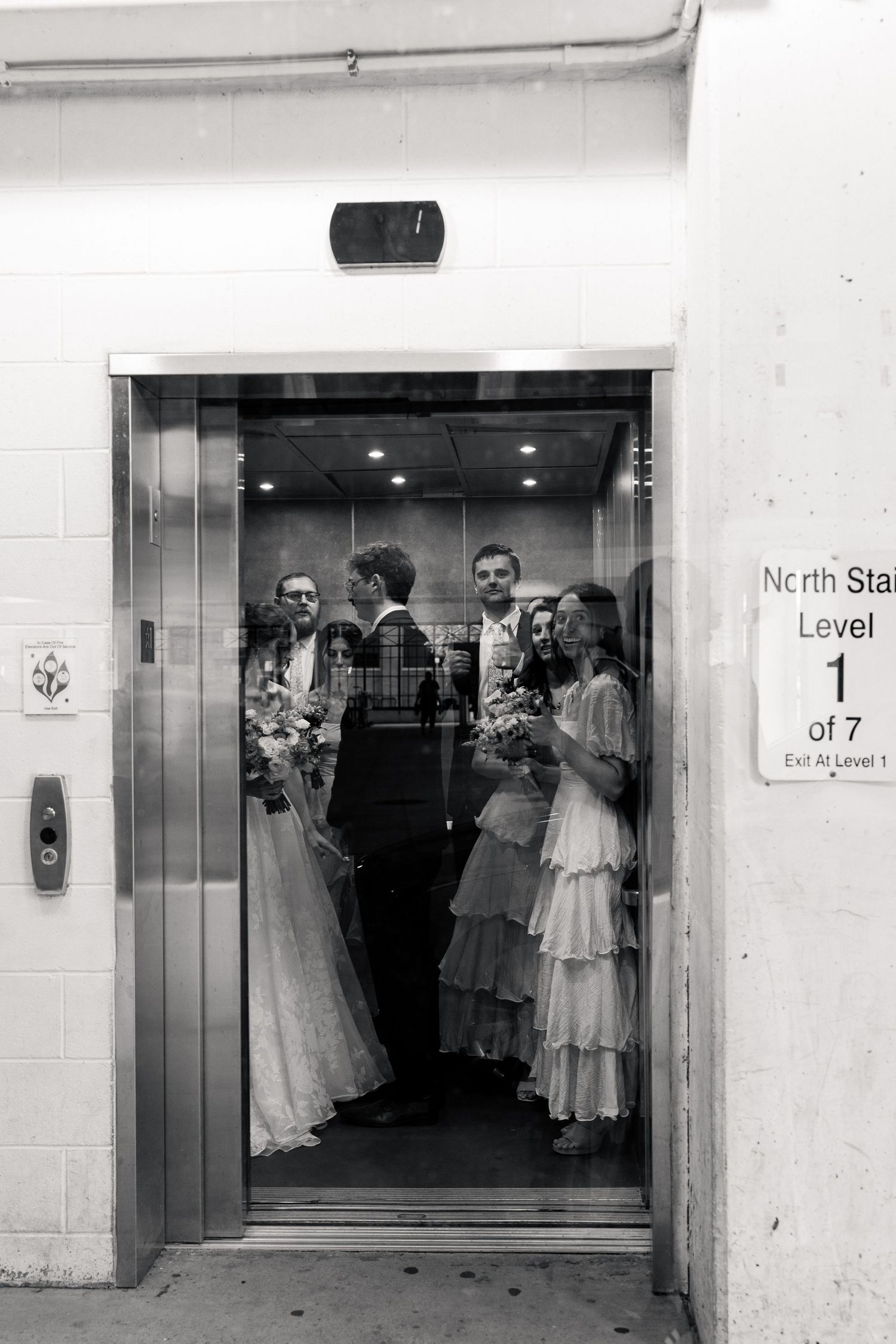 Wedding party riding elevator together in black and white photo at parking garage.