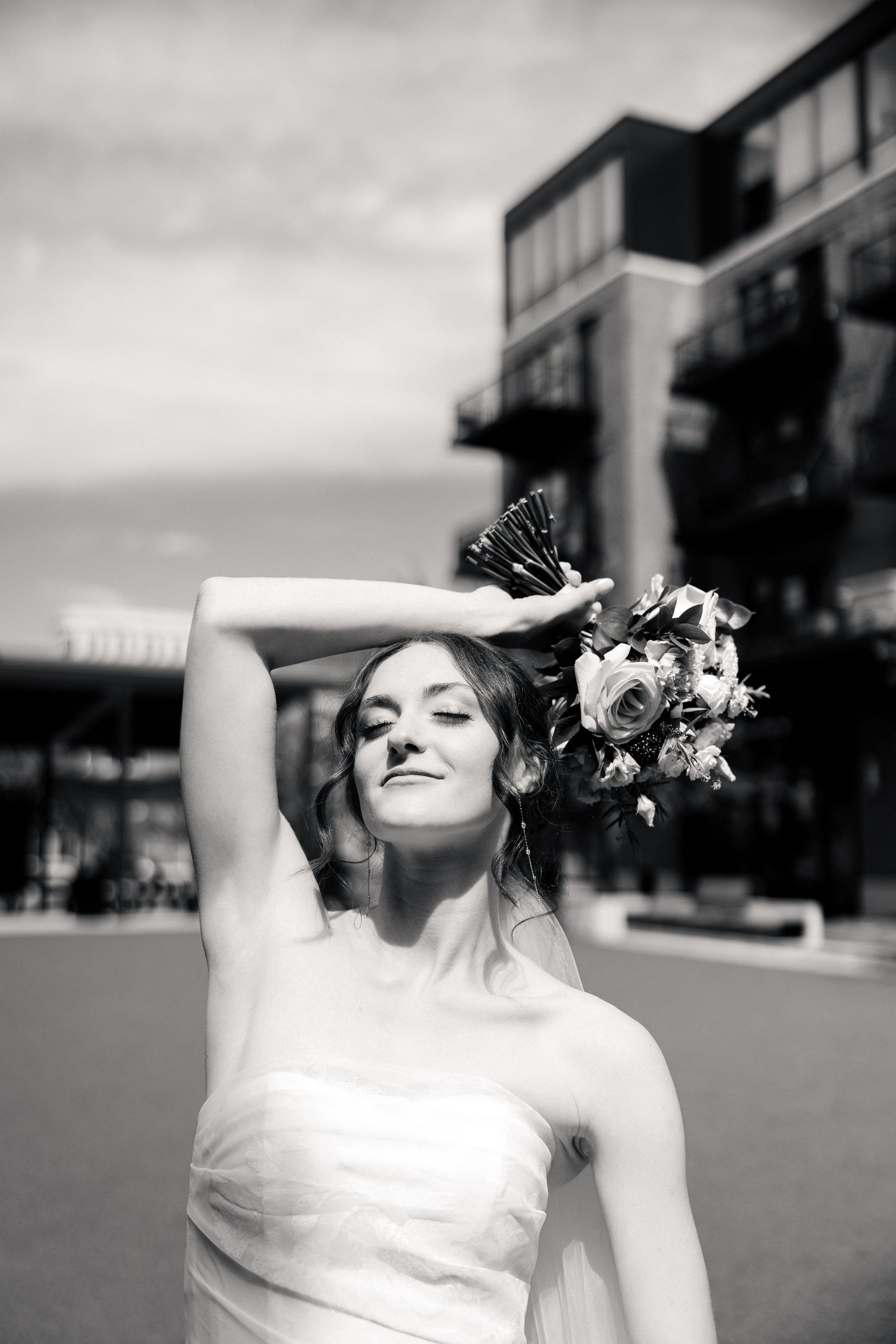 Black and white portrait of a bride holding bouquet overhead with eyes closed outdoors.