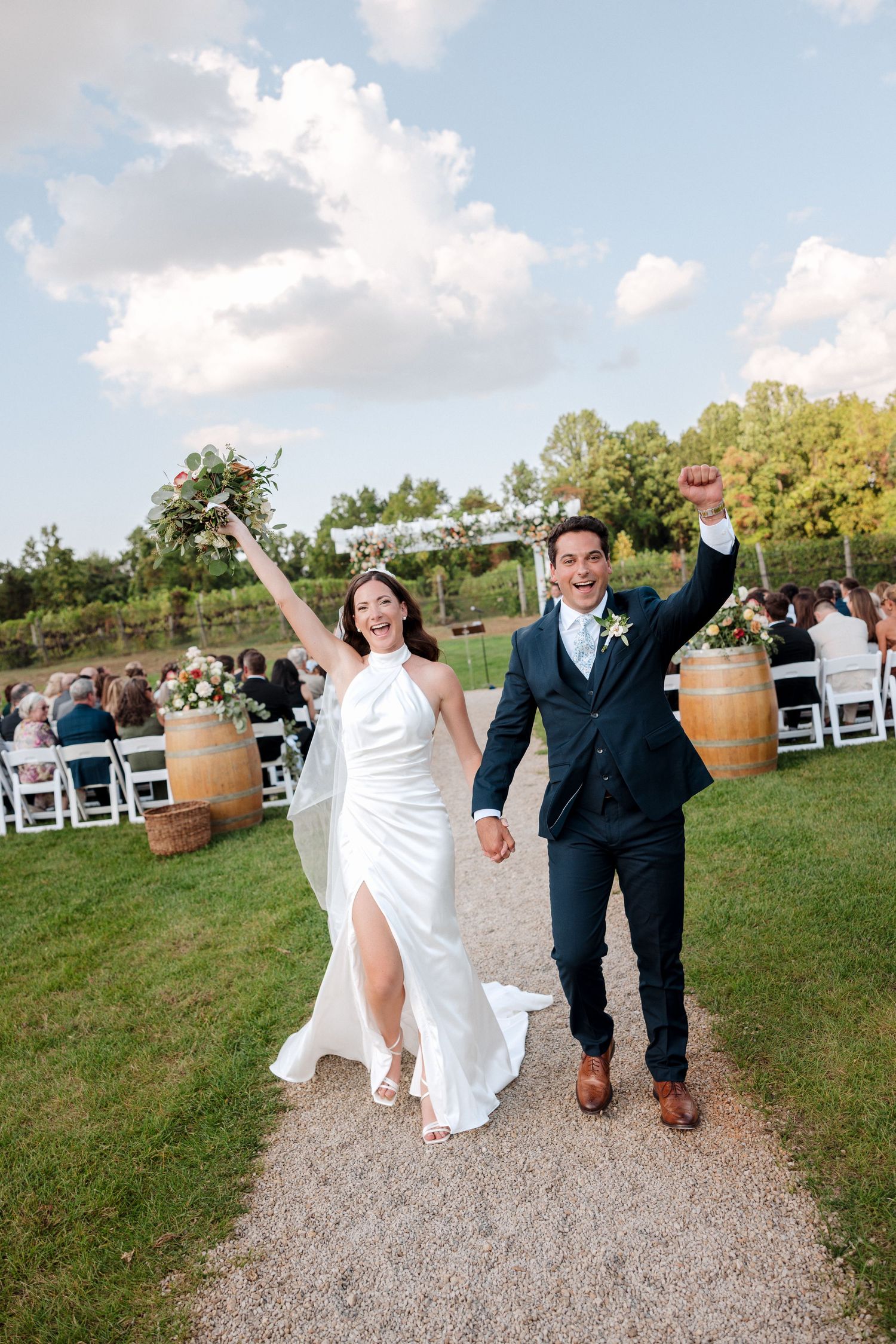 Joyful newlyweds celebrate after outdoor vineyard wedding ceremony, bride in white halter gown, groom in navy suit, arms raised.