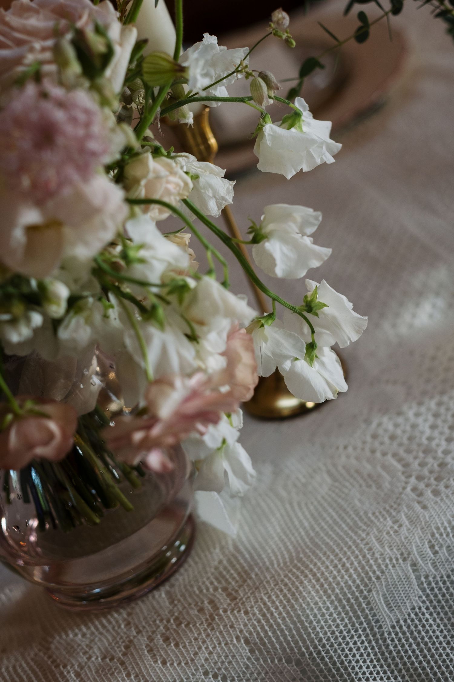 Delicate white and pink sweet pea floral arrangement in glass vase on lace tablecloth.