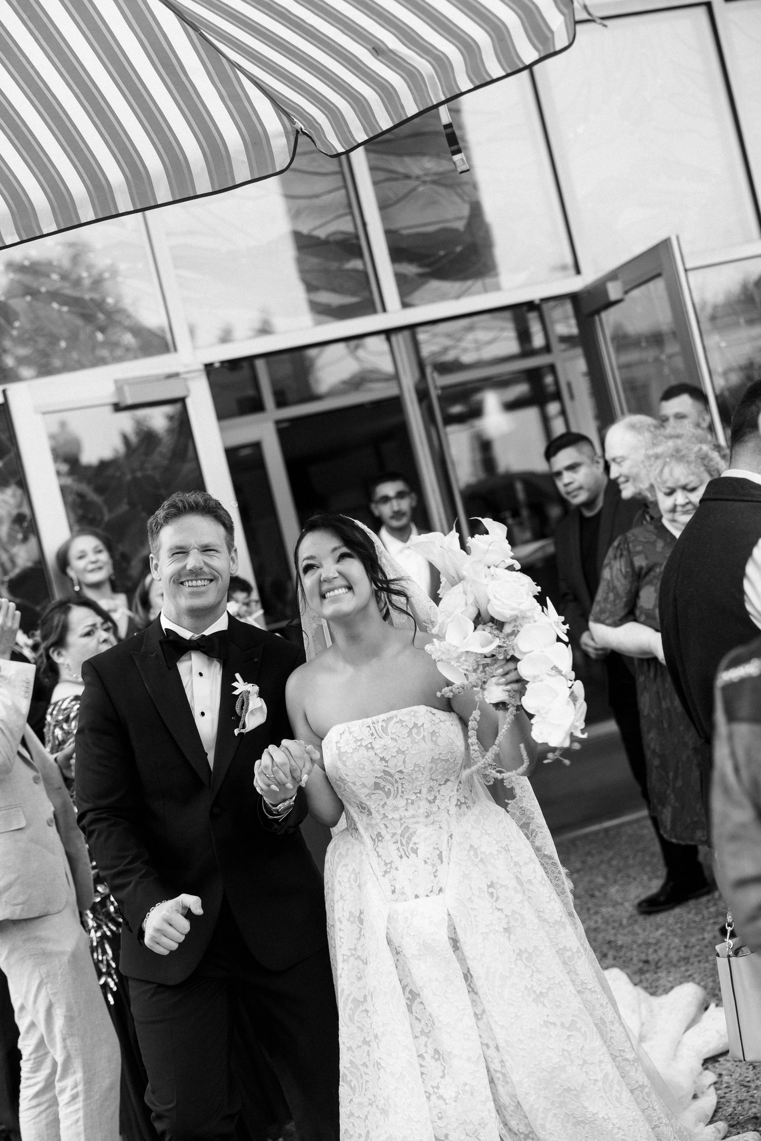 Black and white photo of a smiling bride and groom exiting their wedding venue, surrounded by cheering guests.