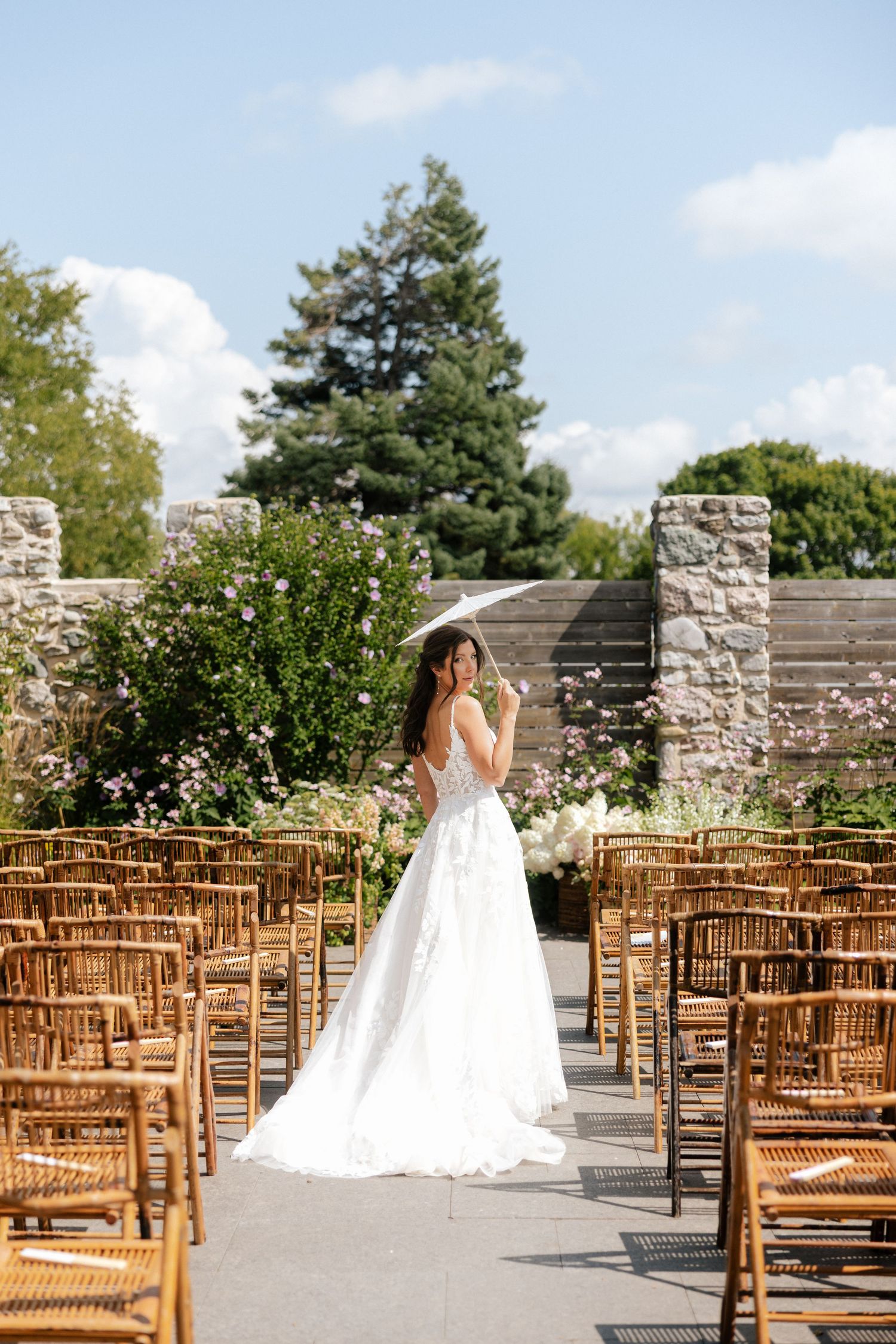 Bride in elegant white ball gown holding a parasol walks down the aisle of an outdoor garden wedding ceremony.