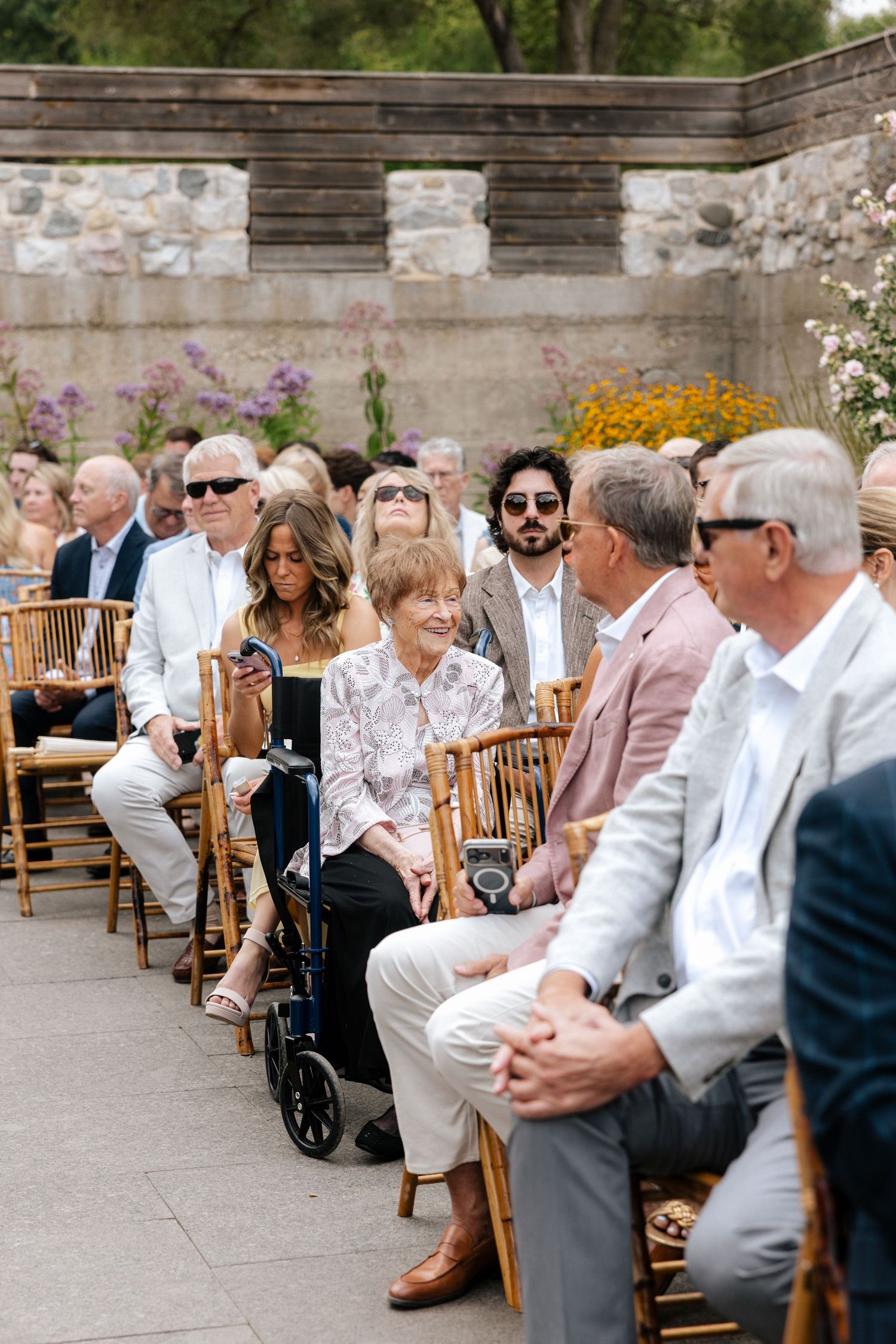 Wedding guests seated in bamboo chairs at outdoor garden ceremony on sunny day.