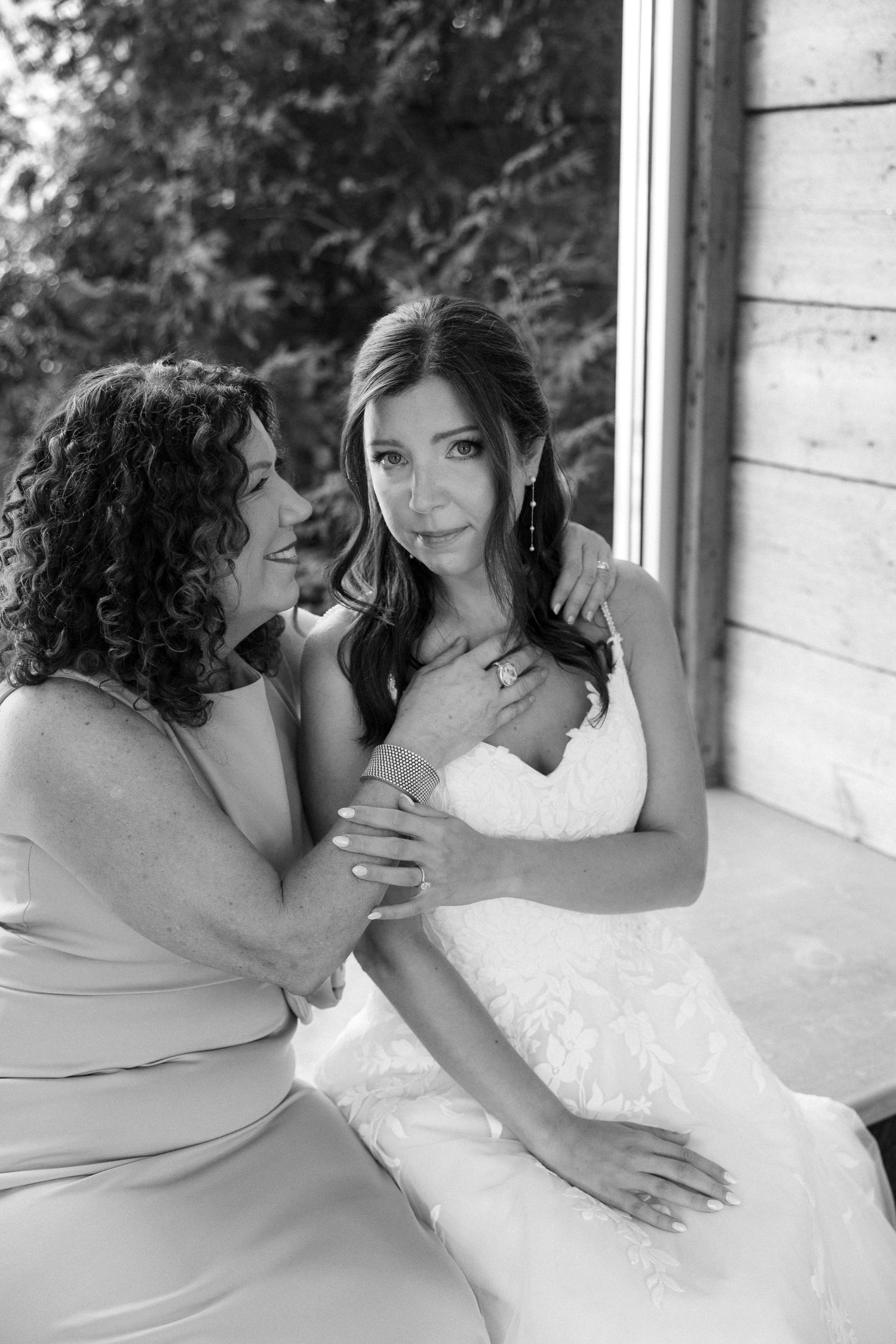 Black and white photo of a bride in white gown sharing a tender moment with a woman in a light dress near a window.