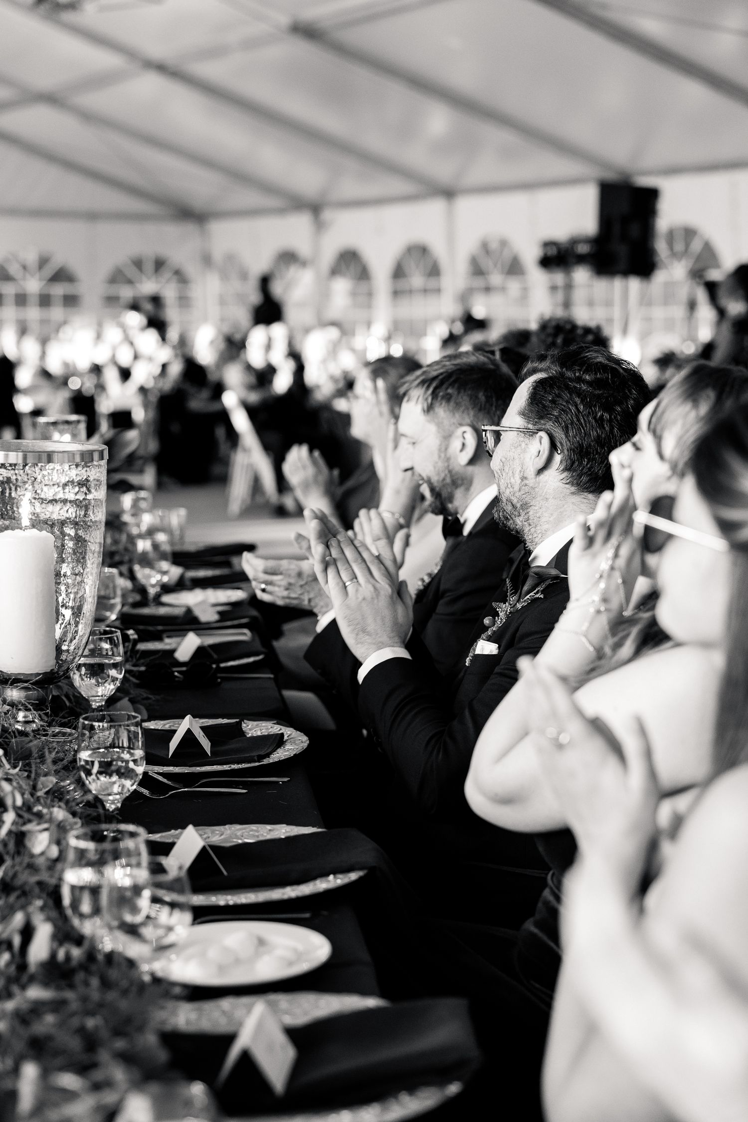 Black and white photo of wedding guests clapping at a formal reception dinner table under a white tent.