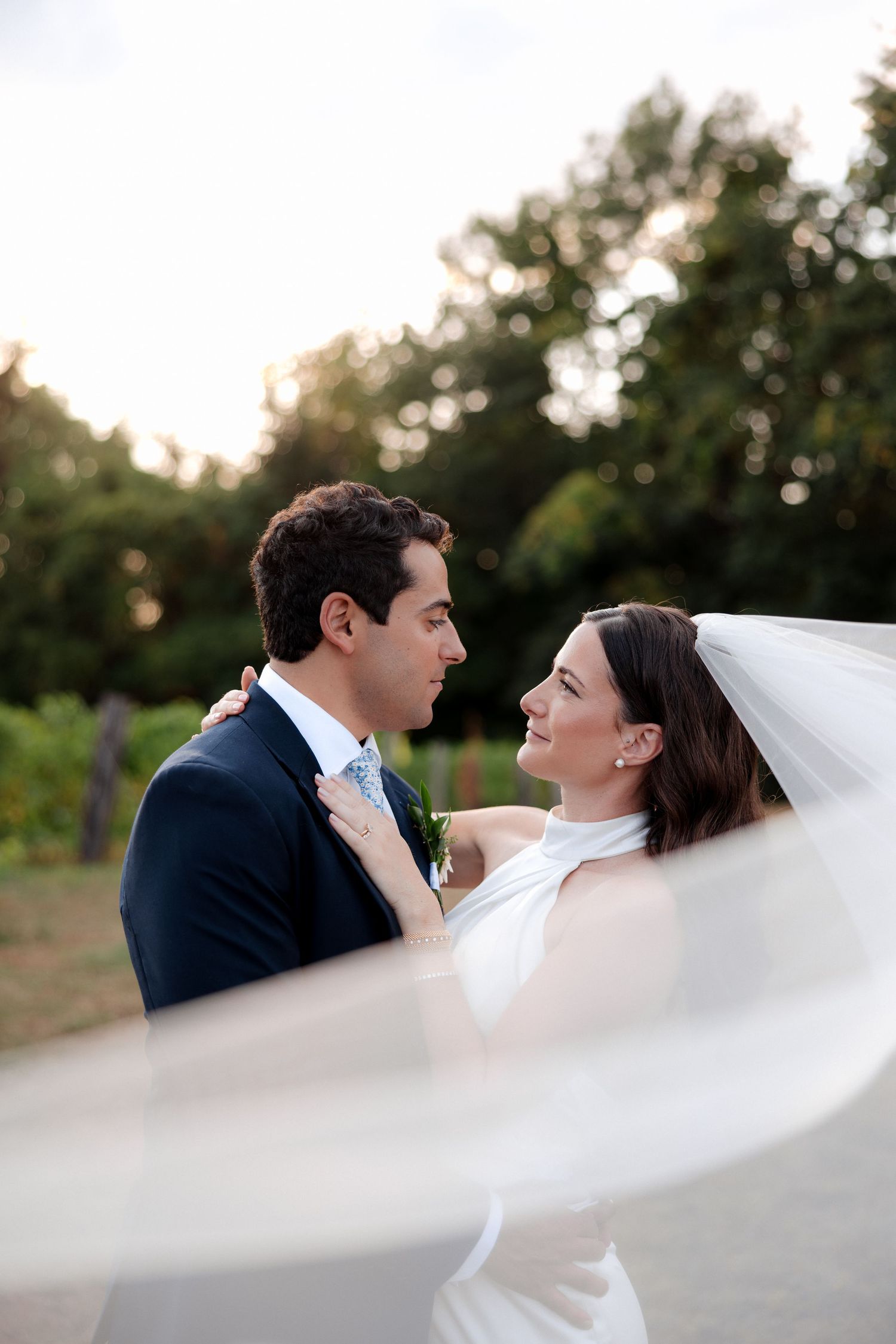 Bride and groom share a romantic moment outdoors, her veil flowing, surrounded by lush greenery at golden hour.