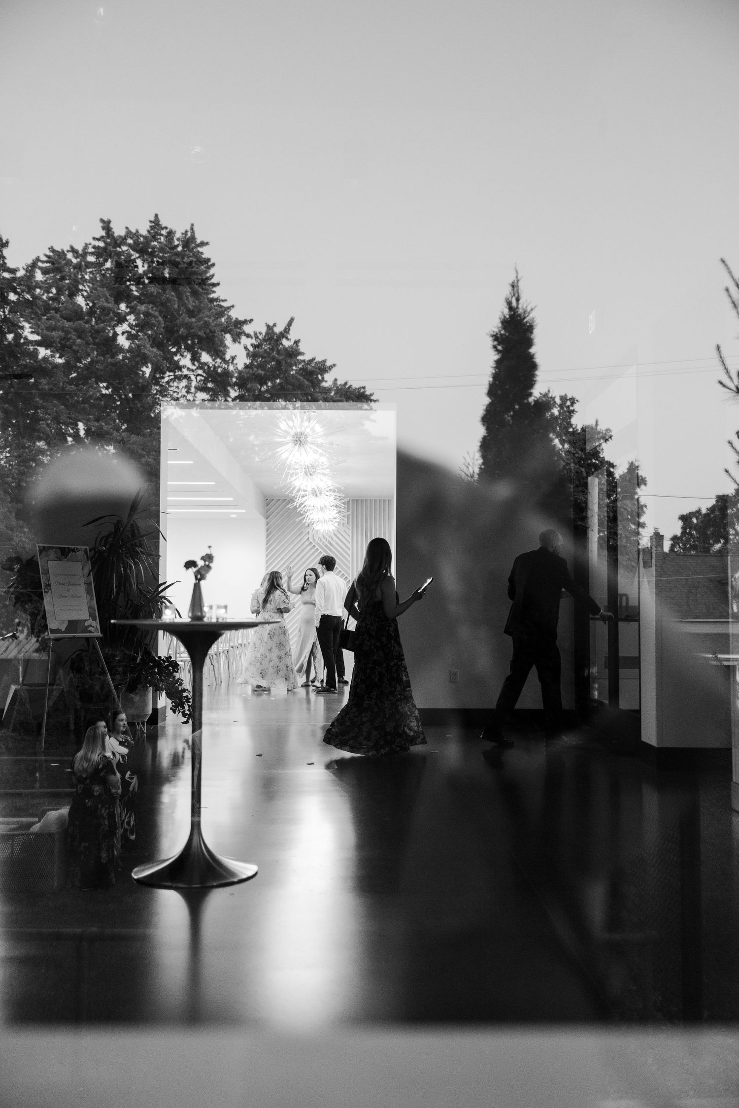 Black and white photo of elegant wedding reception through glass, guests mingling near floral arrangements and tall cocktail tables.