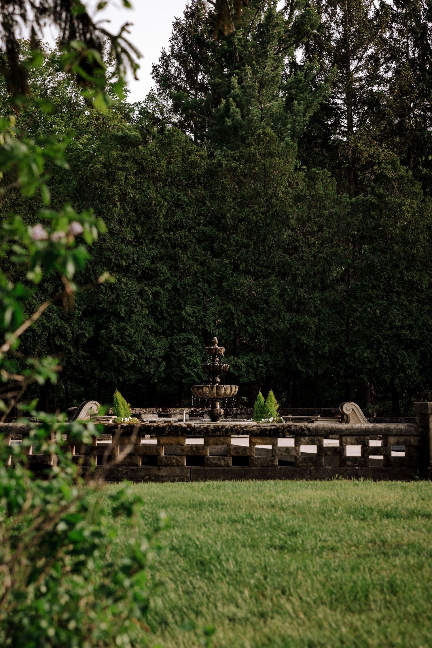Stone garden fountain surrounded by lush green trees and manicured lawn in a serene outdoor setting.